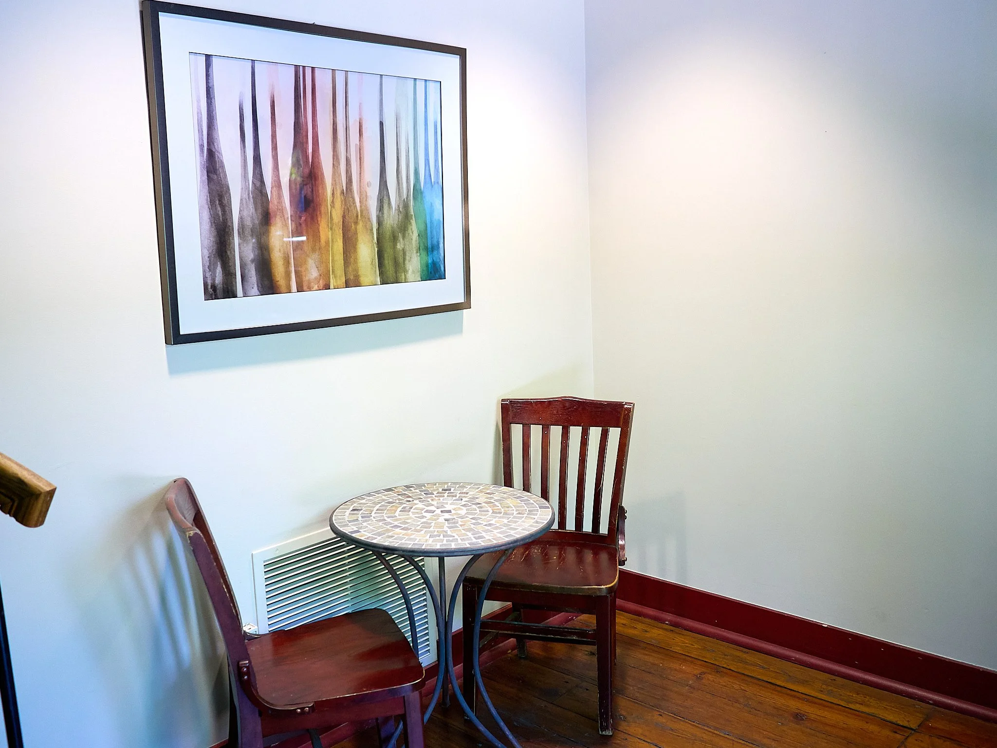 A small table with a mosaic top surrounded by two wooden chairs with slatted backs, positioned against white walls with a framed abstract painting of colorful bottles hanging on the wall, and a hardwood floor.
