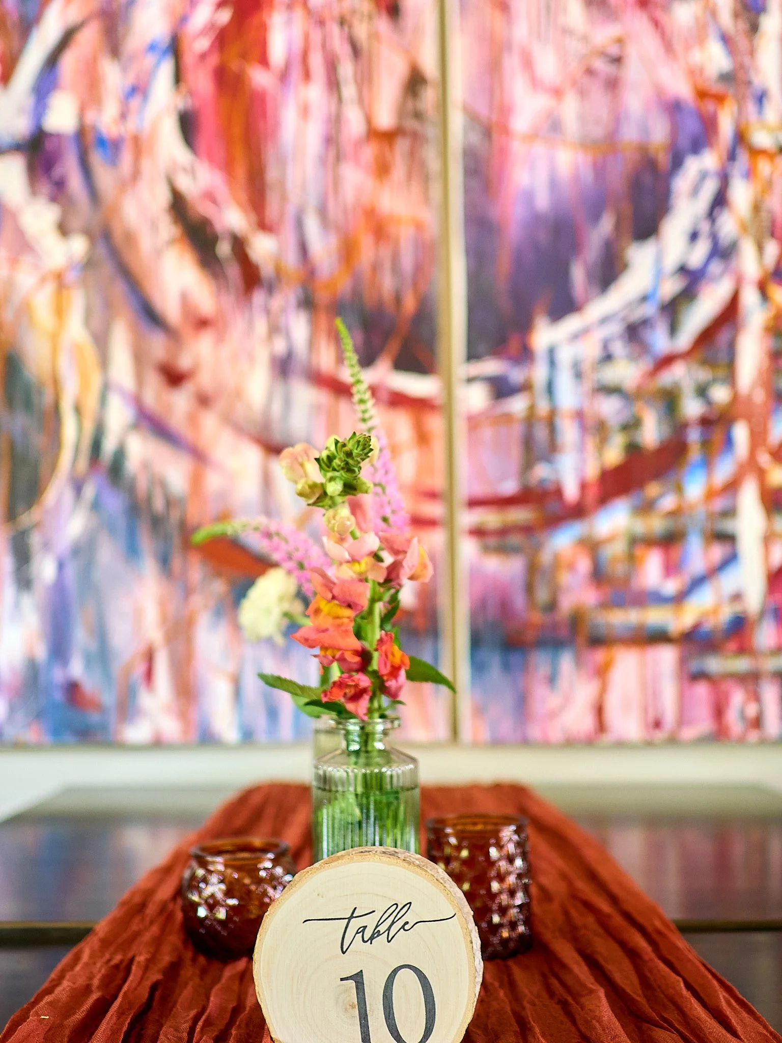 A table centerpiece with a small glass bottle holding pink and green flowers, flanked by two brown decorative glass candle holders, on a rust-colored table runner with a wooden table number sign labeled 'table 10' in front of an abstract pink and pur