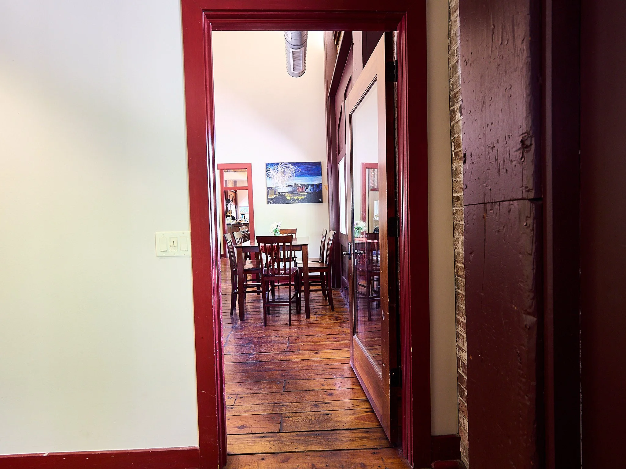 An interior view of a dining area with a wooden table and chairs, artwork on the wall, and a doorway leading to another room.