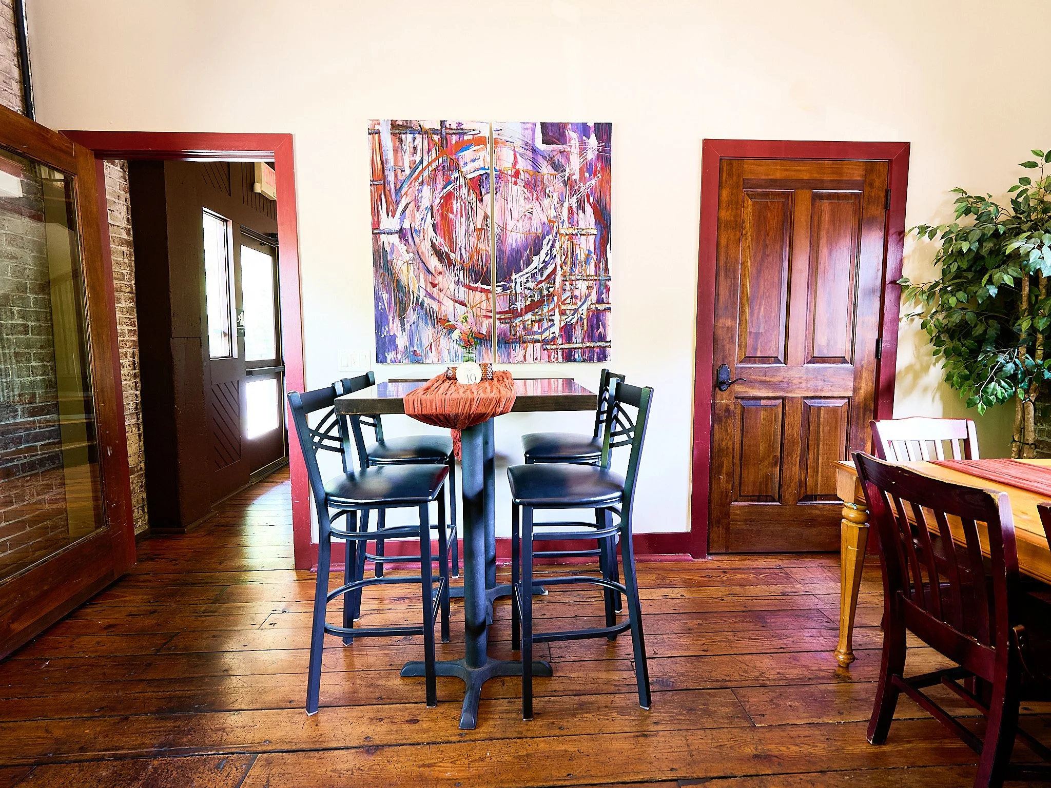 Interior of a dining room with wooden floors, a tall square table with five black chairs, a large colorful abstract painting on the wall, a wooden door, a potted plant, and part of another table with chairs.