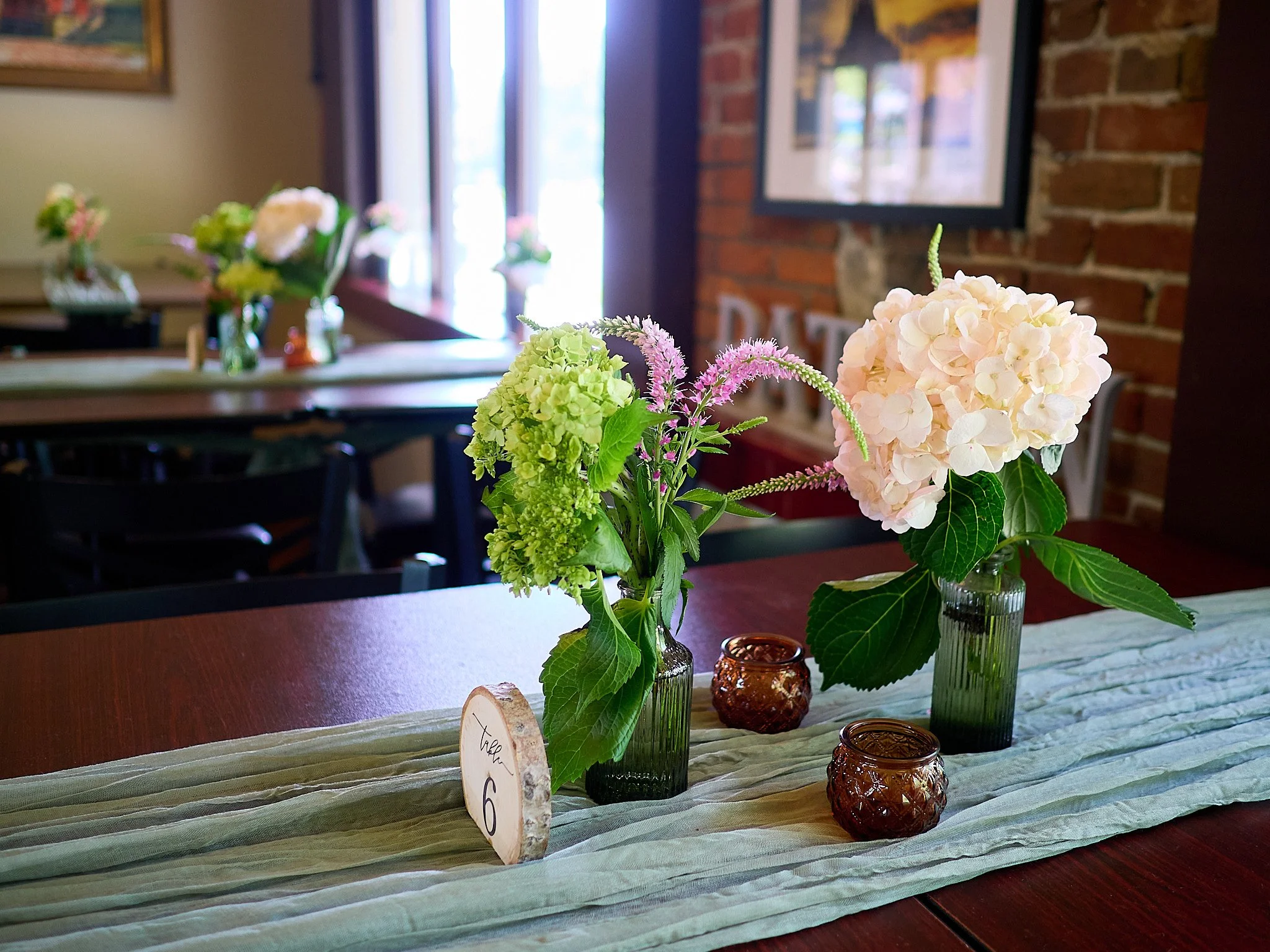 Table centerpiece with pink and green hydrangea flowers in glass vases, wooden table number, and amber candle holders on a light-colored table runner in a cozy restaurant setting.