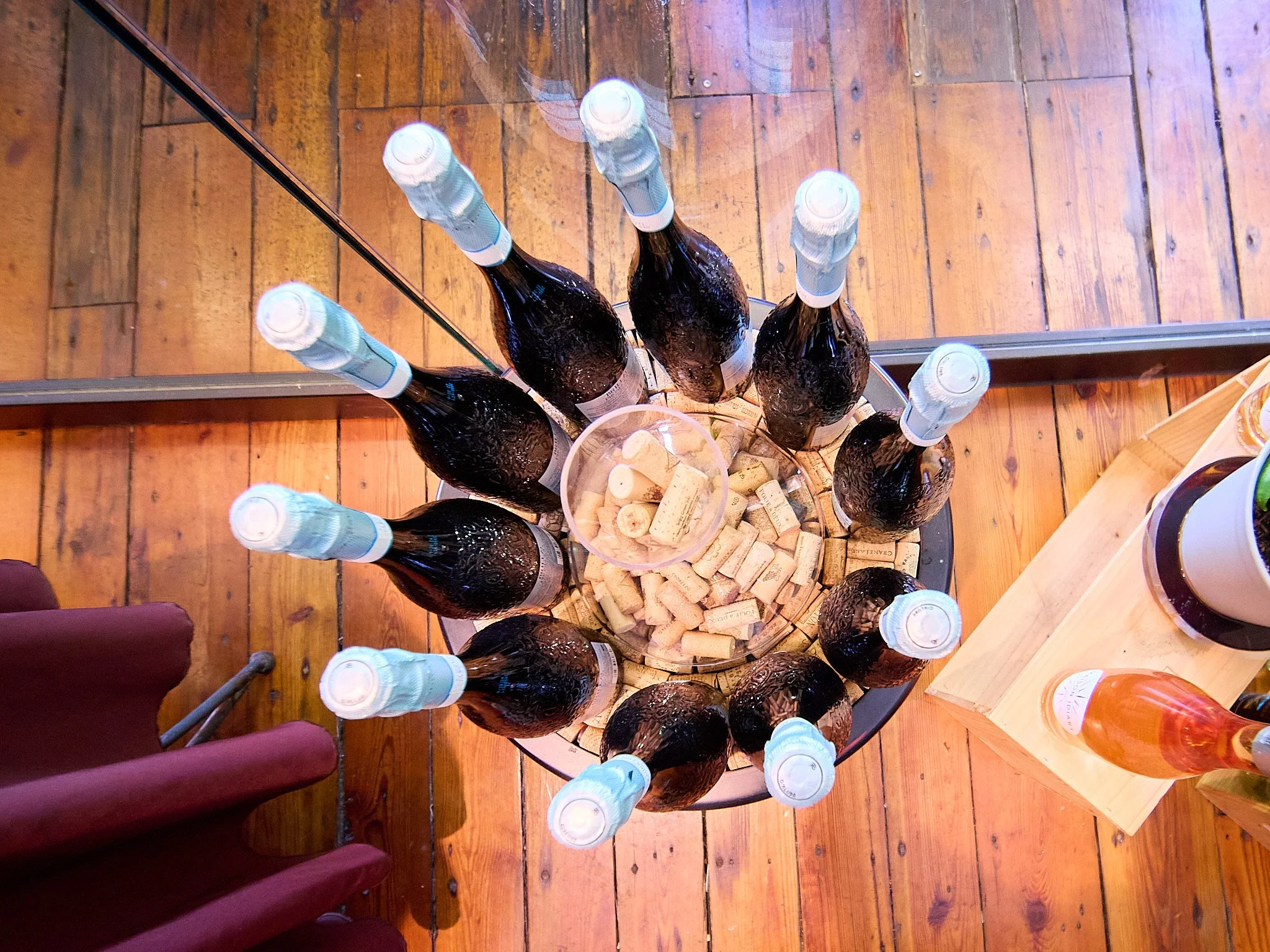 A top-down view of a display with multiple bottles of beer arranged in a circle around a central transparent container filled with wine corks. The display rests on a wooden floor, with part of a glass door or window reflecting the scene.
