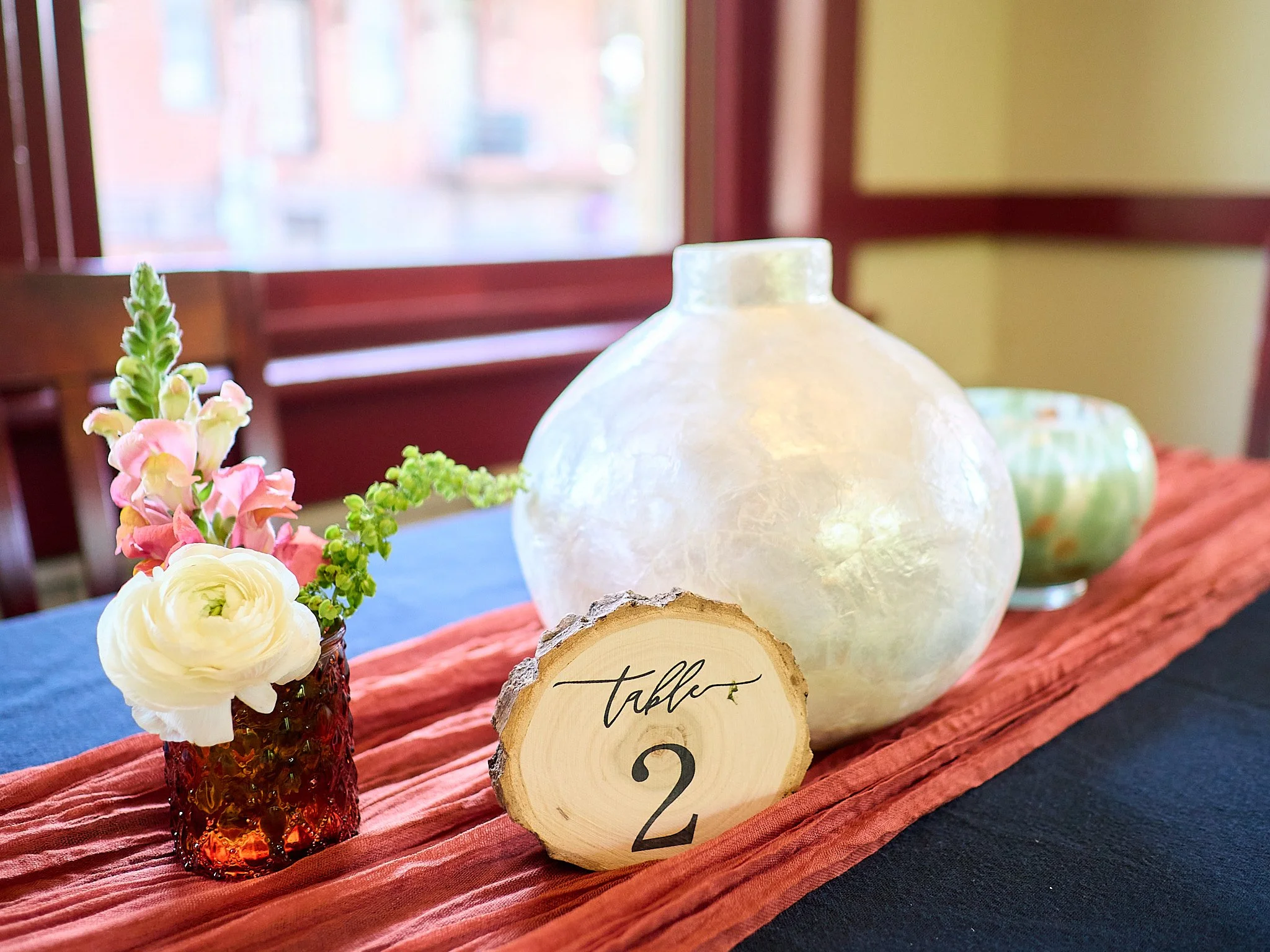 Decorative table setting with a small vase of pink and white flowers, a large white vase, a green vase, and a wooden table number sign with the number 2, on a fabric table runner in a warmly lit room.