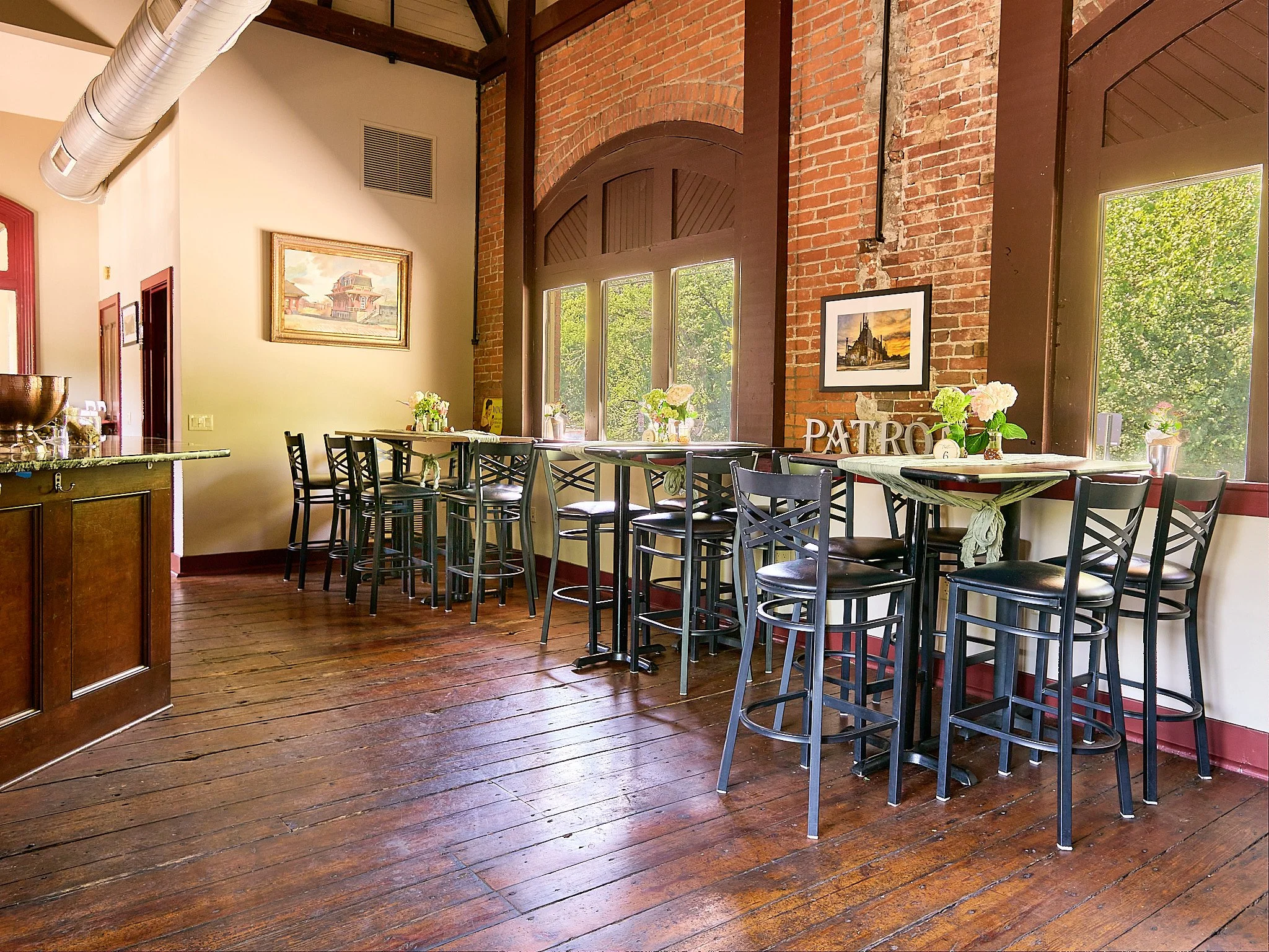 Interior of a cafe or restaurant with wooden floors, a high brick and wood wall with large windows, and high tables with black chairs. Decor includes framed pictures, vases of flowers, and a decorative letter sign spelling 'PATRO'.