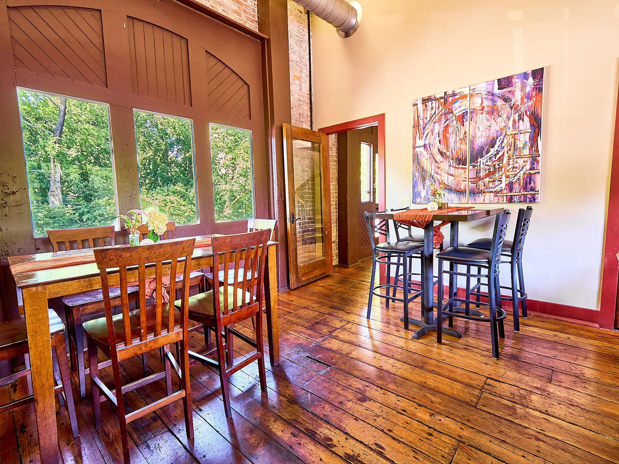 Cozy cafe interior with wooden tables and chairs, large window with outdoor greenery, colorful abstract painting on the wall, and hardwood floors.