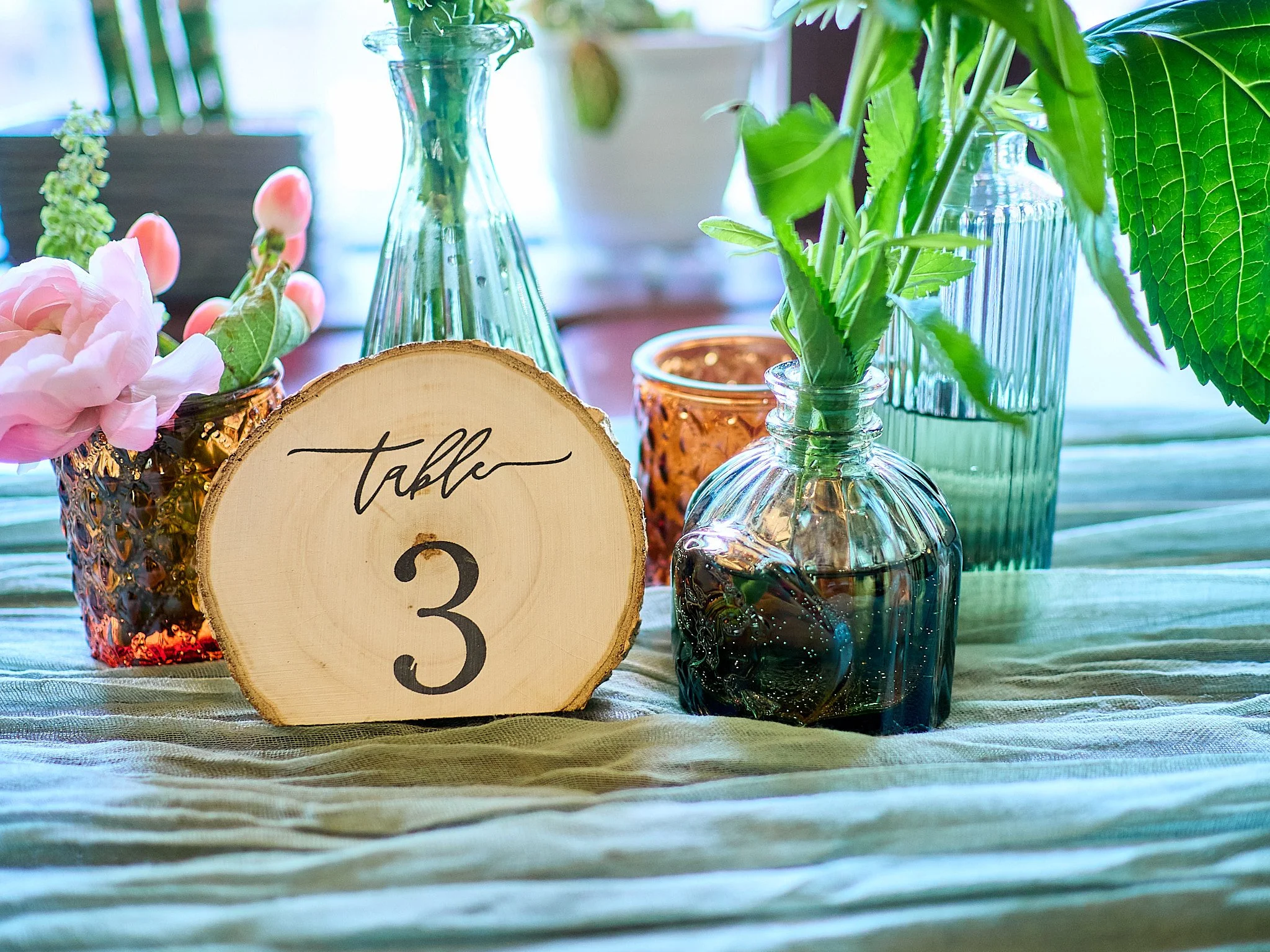 Table centerpiece with glass bottles and a wooden table number sign reading 'table 3' surrounded by flowers and greenery.