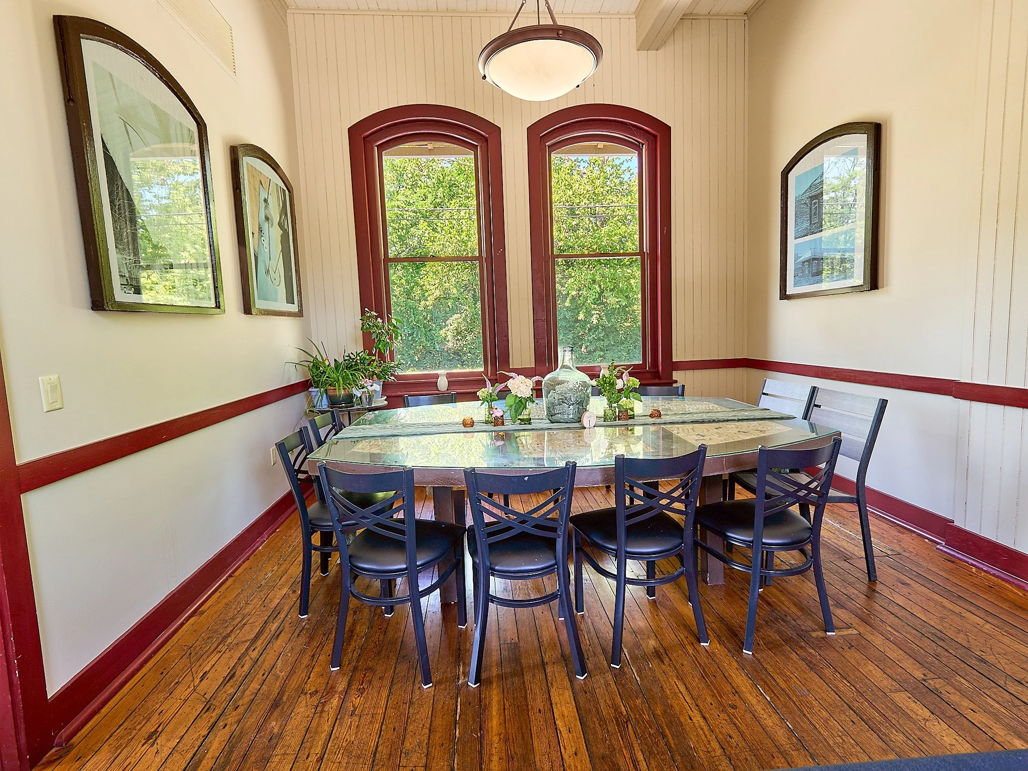 A dining room with a wooden floor, white walls with red trim, and a large window with arched top. There is a round table with a glass top, decorated with flowers and a large glass vessel, and surrounded by black chairs with padded seats. Framed pictu