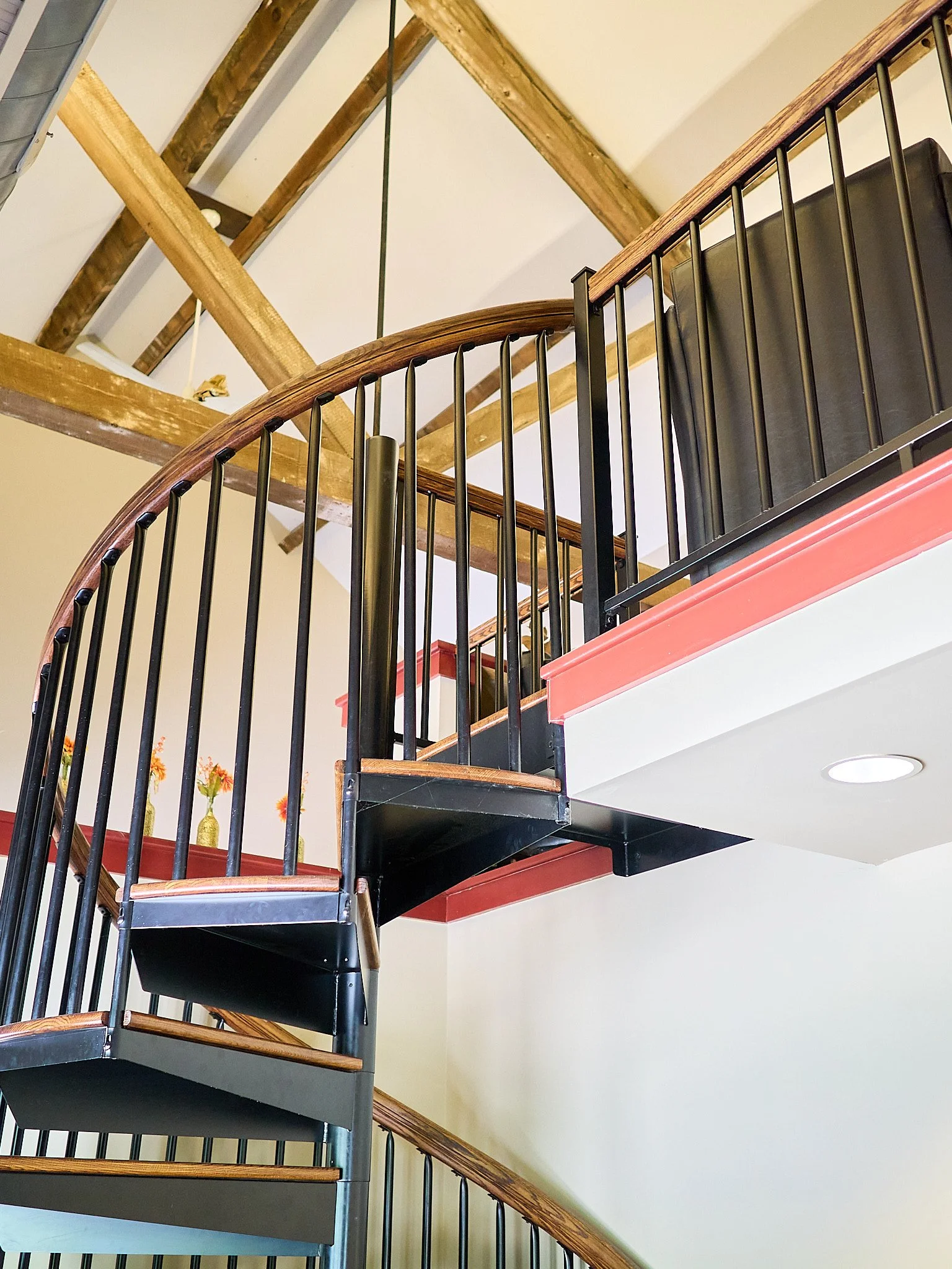 Interior view of a spiral staircase with wooden handrails and black metal balusters, leading to an upper floor with exposed wooden beams on the ceiling.