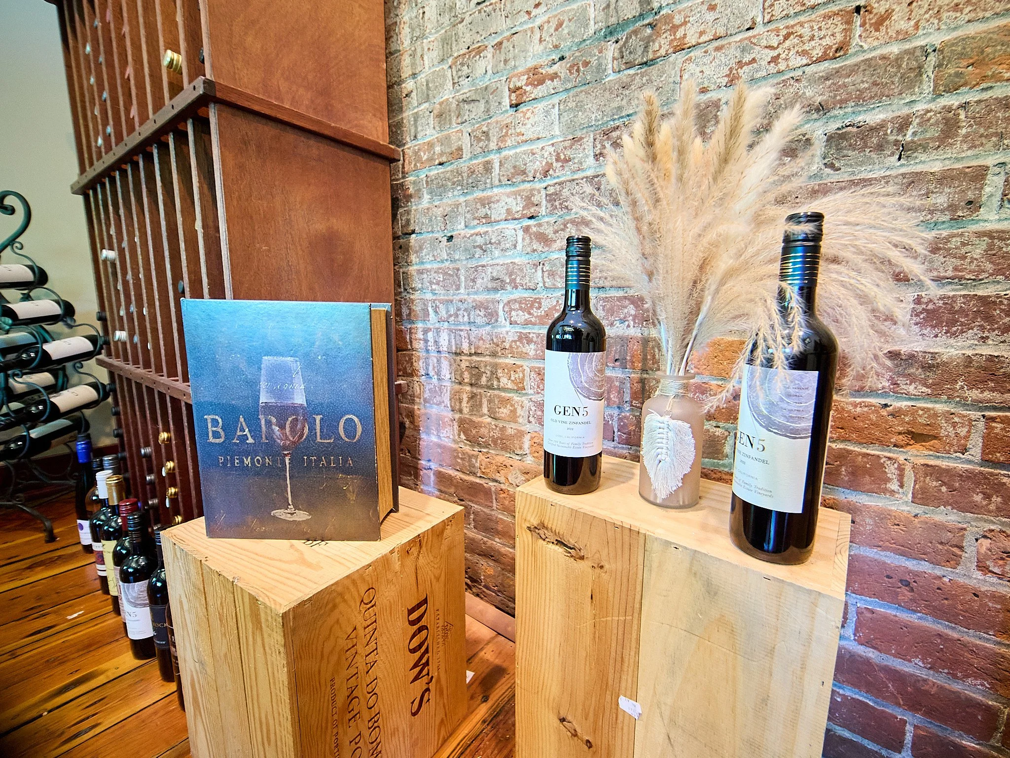 Display of wine bottles on wooden pedestals against a brick wall, with decorative dried grasses in a vase and a wine-related book.