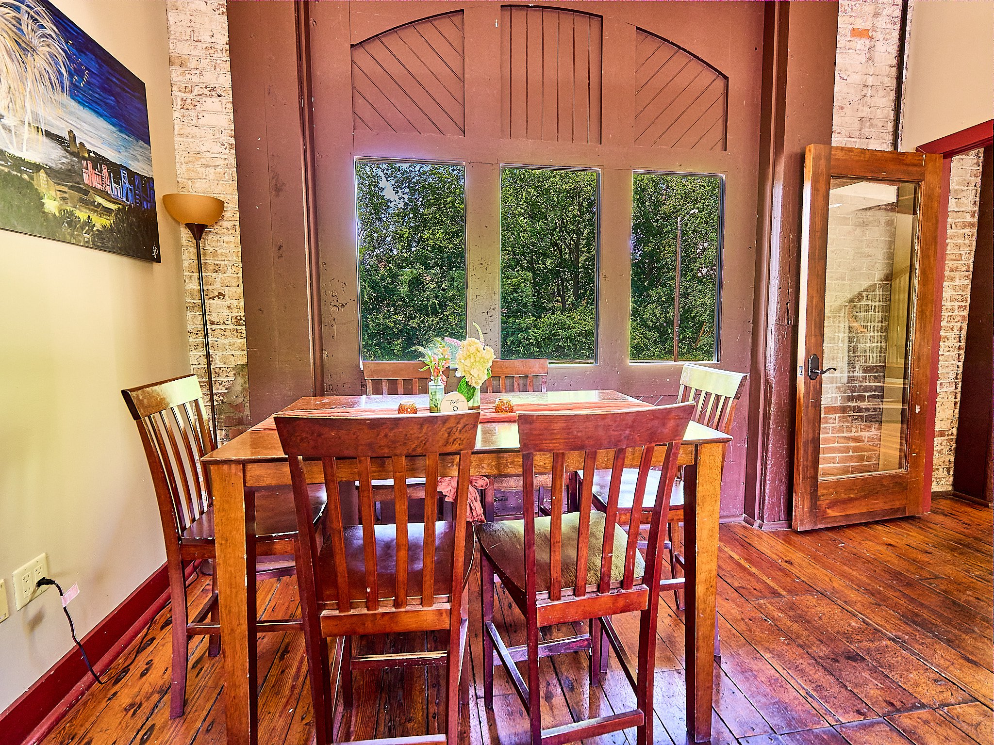 Wooden dining table with four chairs in front of windows showing green trees, next to a brick wall and a glass door.