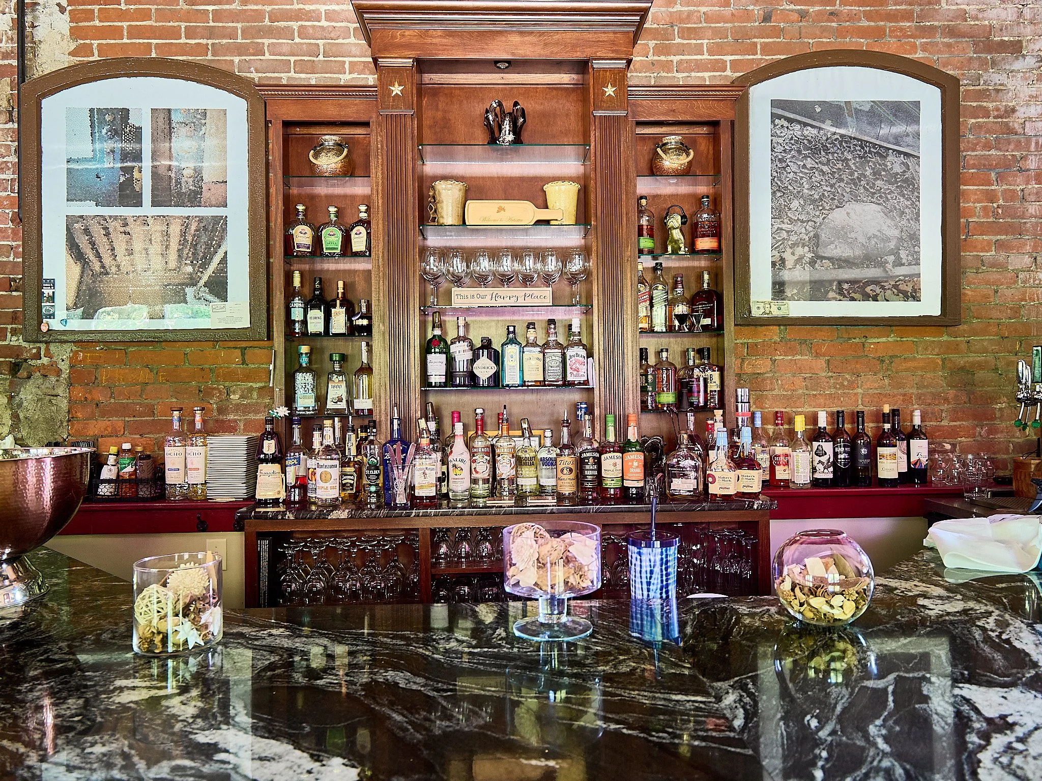 A bar with a variety of liquor bottles on shelves, a dark marble countertop with decorative glass jars, and framed wall art on a brick wall.