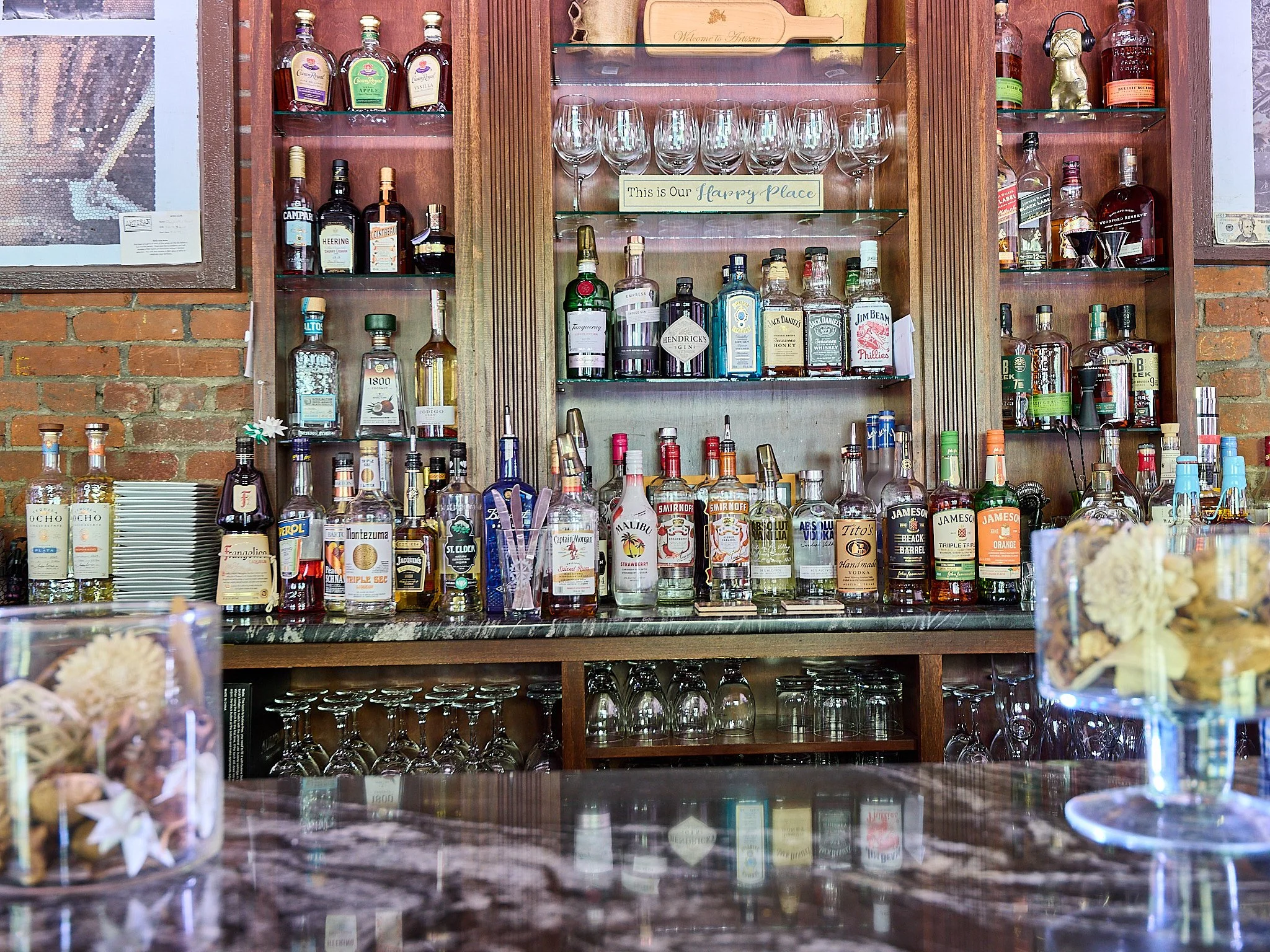 A bar shelf with various bottles of alcohol, including whiskey, vodka, and gin, arranged on glass and wooden shelves against a brick wall. There are empty glasses, a sign that says 'This is Our Happy Place,' and decorative items like flowers and snac