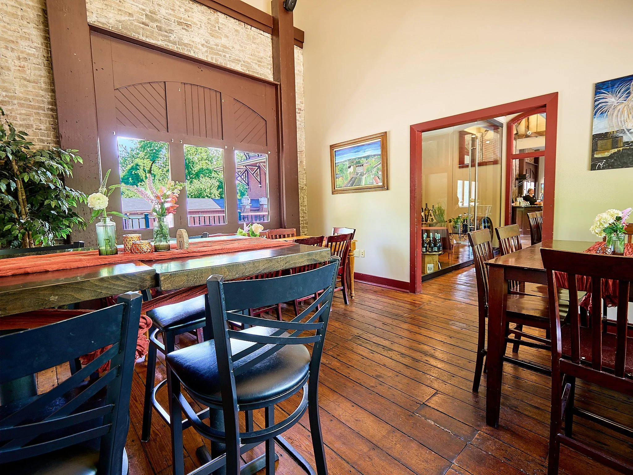 Cozy restaurant interior with wooden floors, a large window, and tables decorated with flowers in vases.