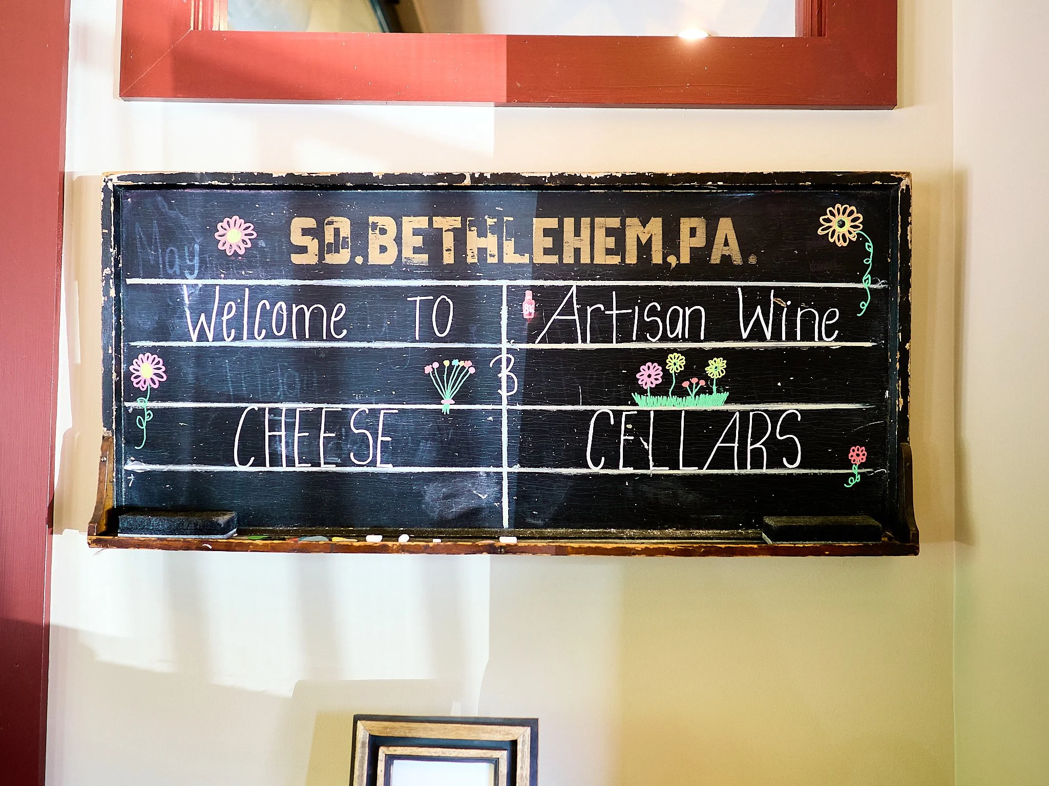 A black chalkboard with colorful flowers and text. The board says, "Welcome to cheese, artisan wine, cellars" and has the date "May" written on the left side.