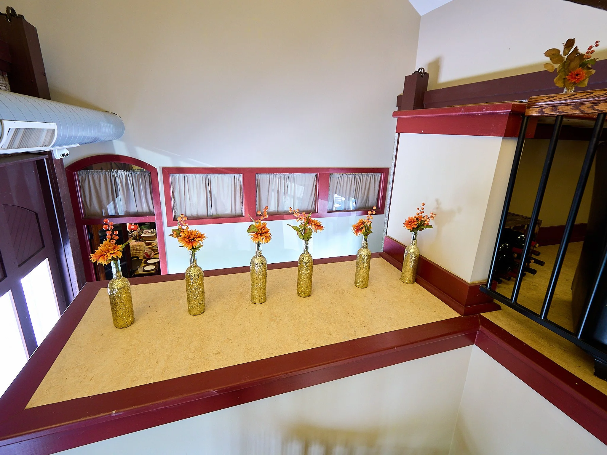 Six gold sparkly vases with orange and yellow flowers on a beige tabletop in a restaurant, with a balcony railing and window in the background.