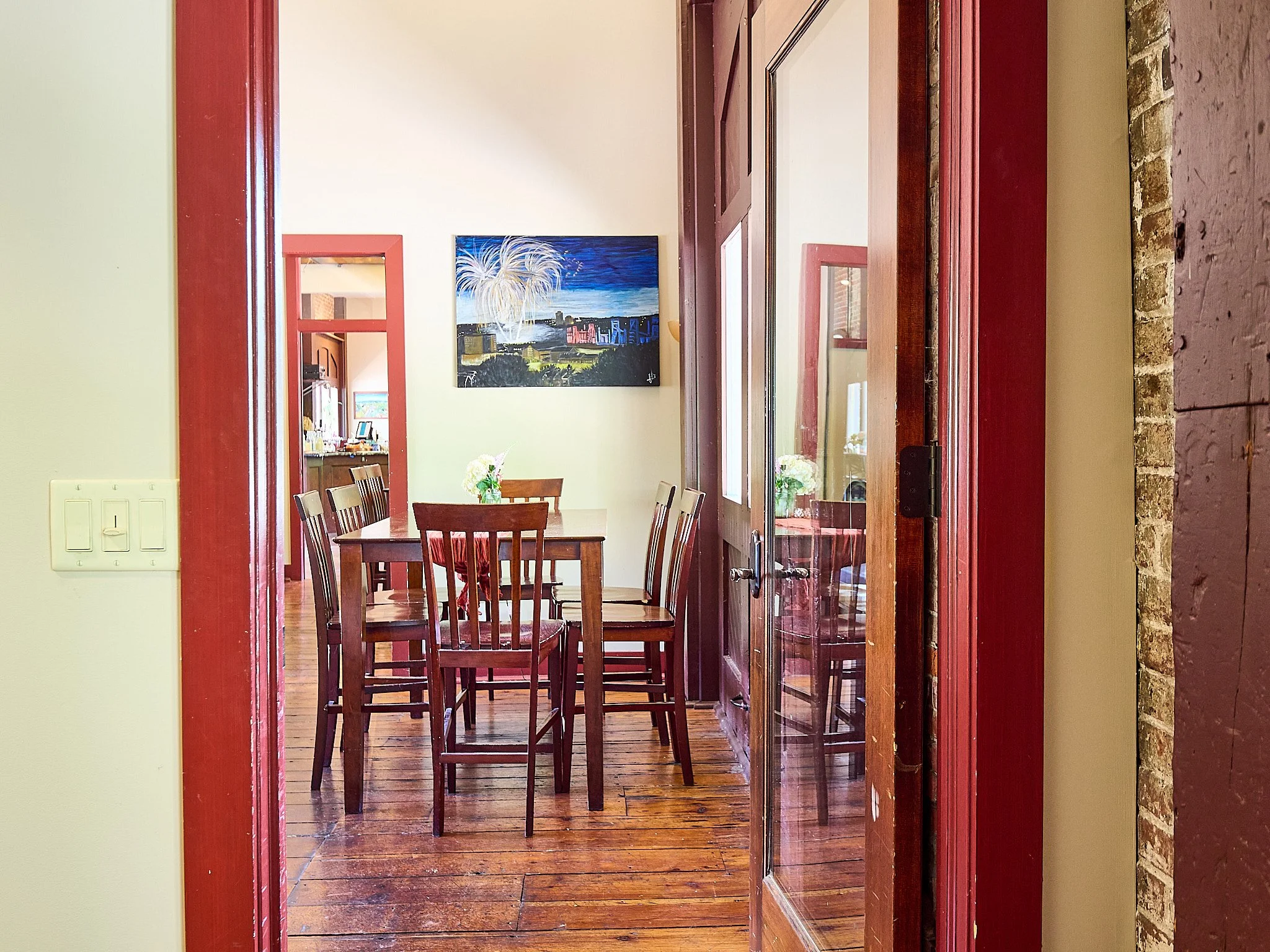 View of a dining room with a wooden table and chairs, a floral decoration, and a painting of fireworks on the wall.