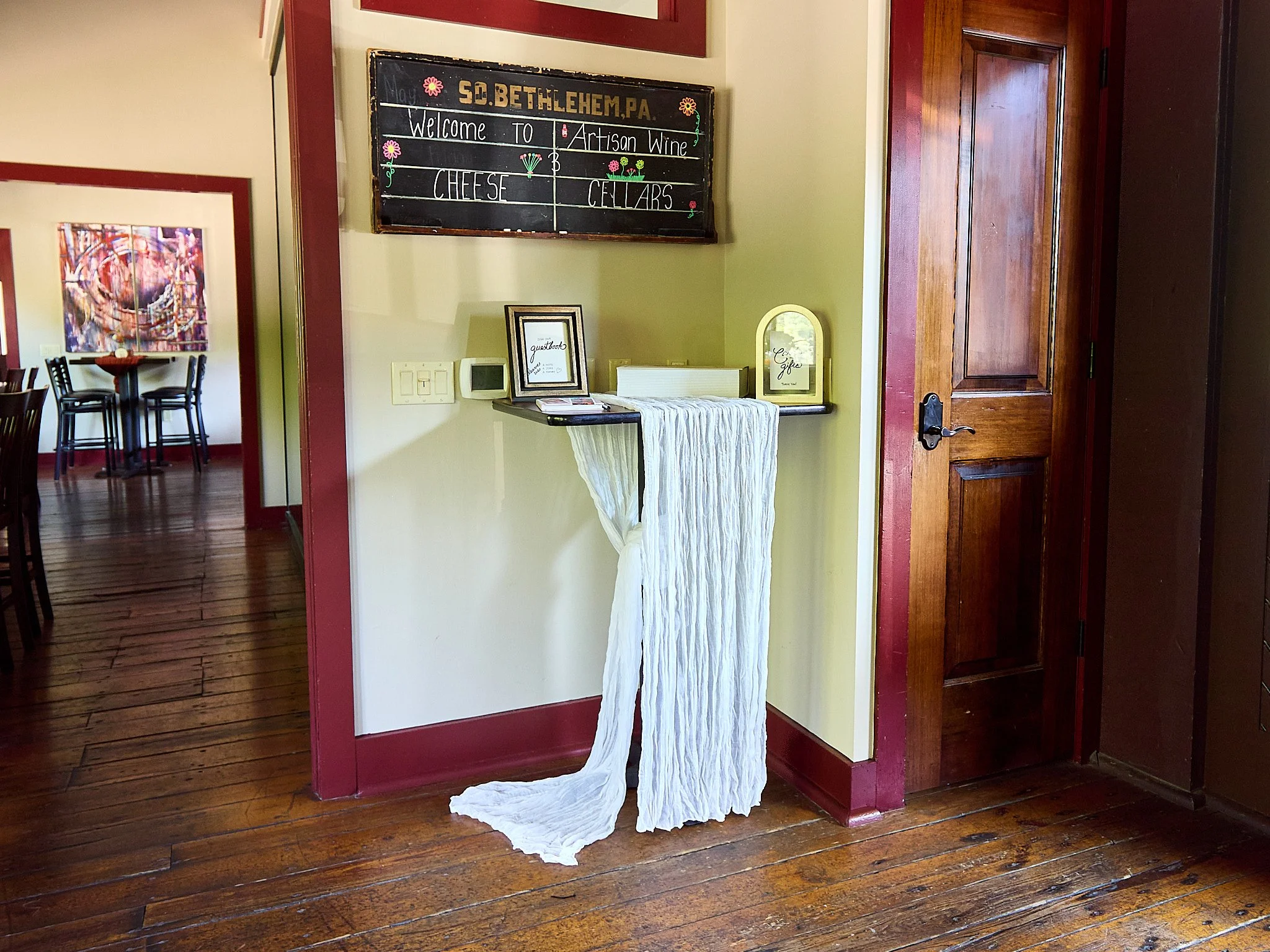 Interior of a restaurant with a blackboard sign above a small floating shelf displaying framed pictures and a white decorative fabric. The blackboard has writing about cheese and wine. To the right, a closed wooden door. The floor is wooden, and ther