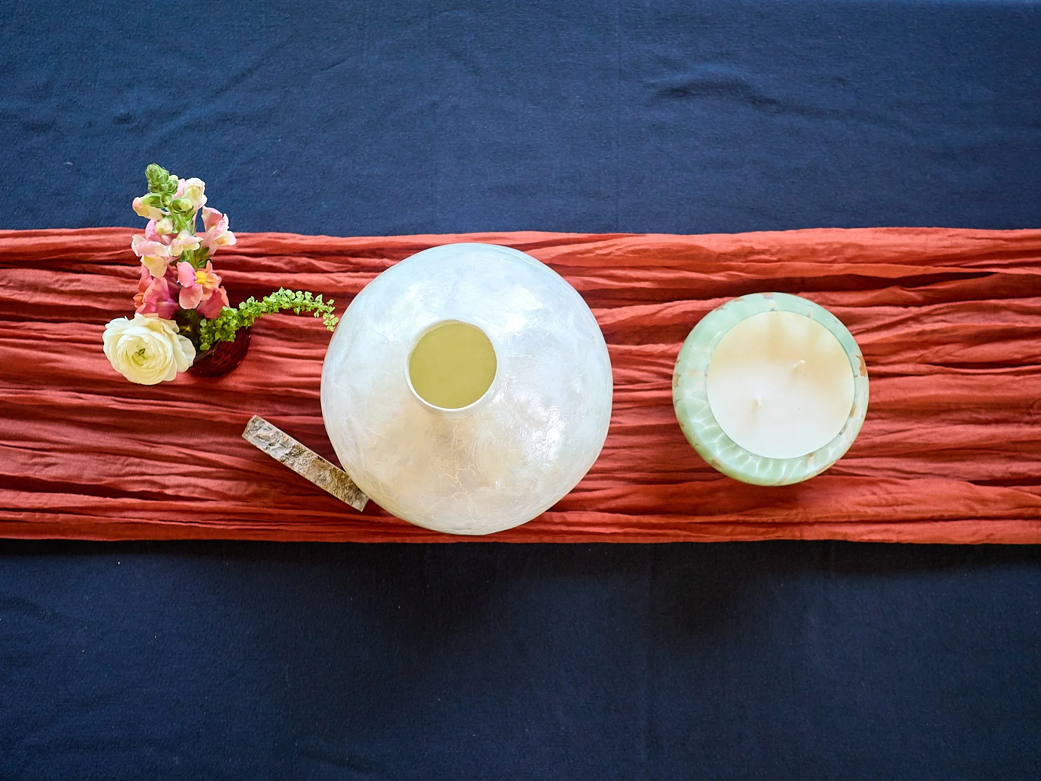 Top-down view of a table arrangement with a small flower bouquet, a spherical white candle holder, and a round green candle on a textured red runner, against a dark blue tablecloth.