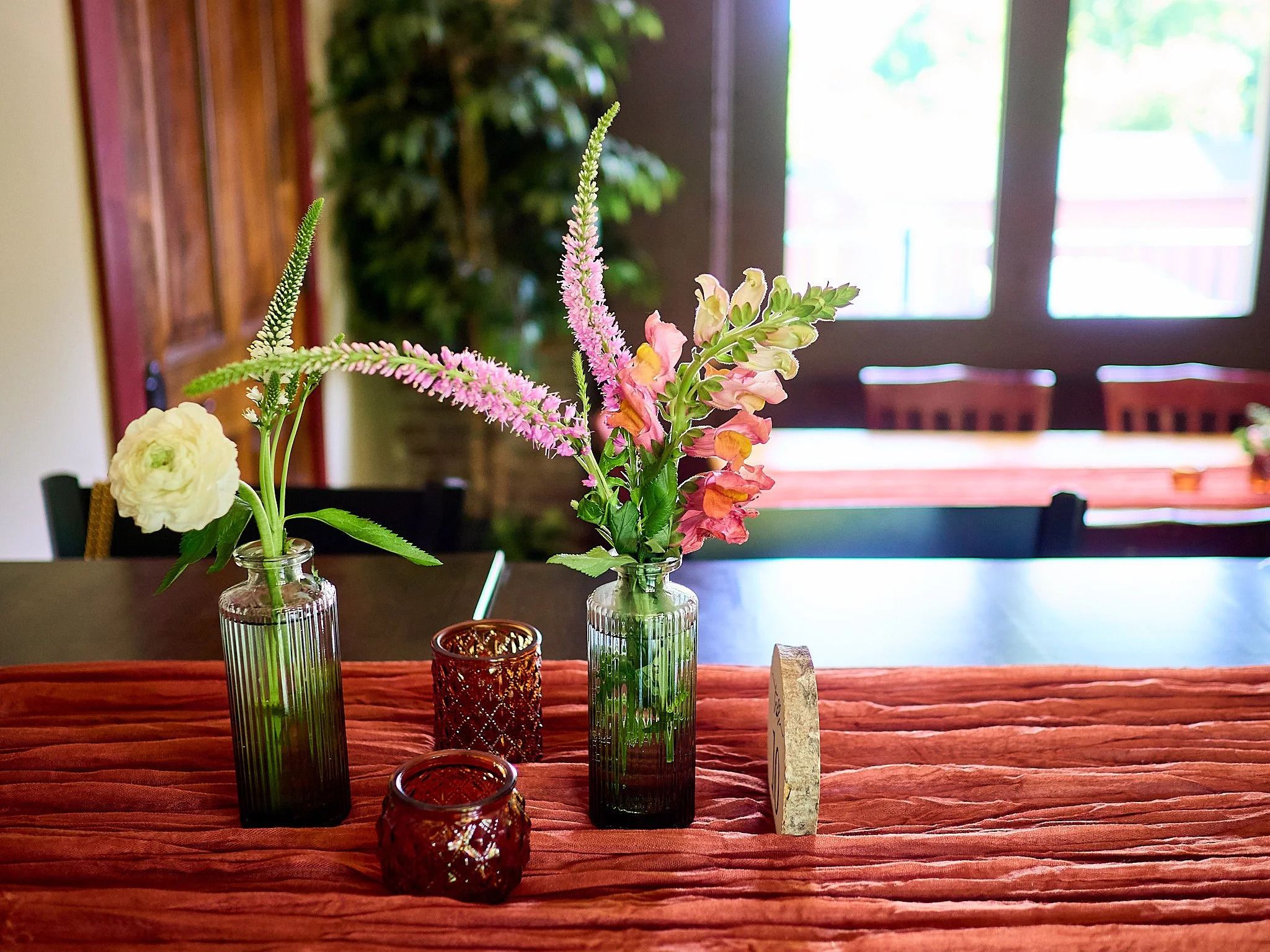 Two glass vases with pink and yellow flowers and greenery, alongside two small amber glass candle holders, on a textured red tablecloth with a dining room in the background.