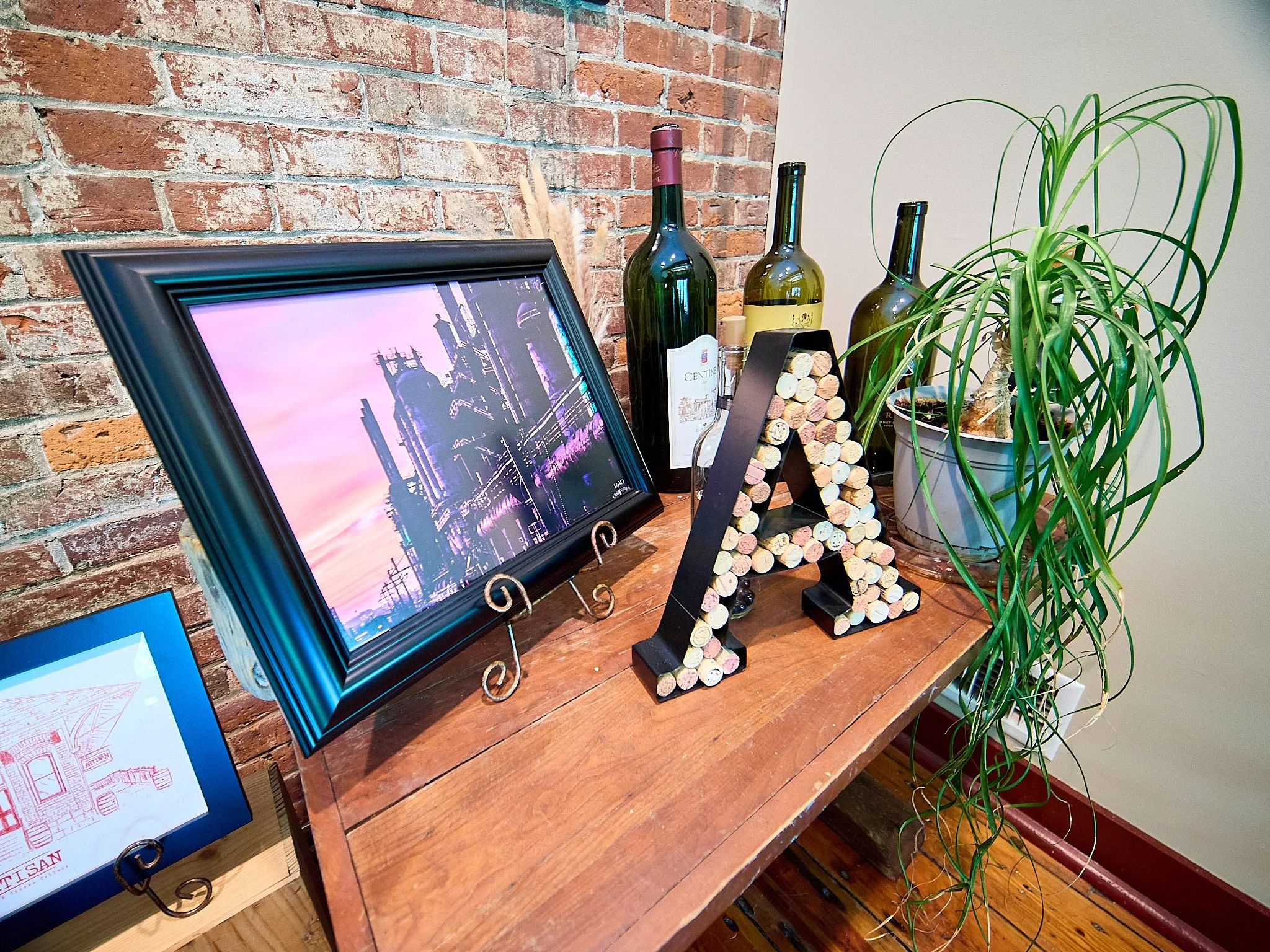Decorative wooden table with a framed photo of a city skyline at sunset, craft beer bottles, a black letter A made of corks, and a large potted spider plant, against a brick wall.