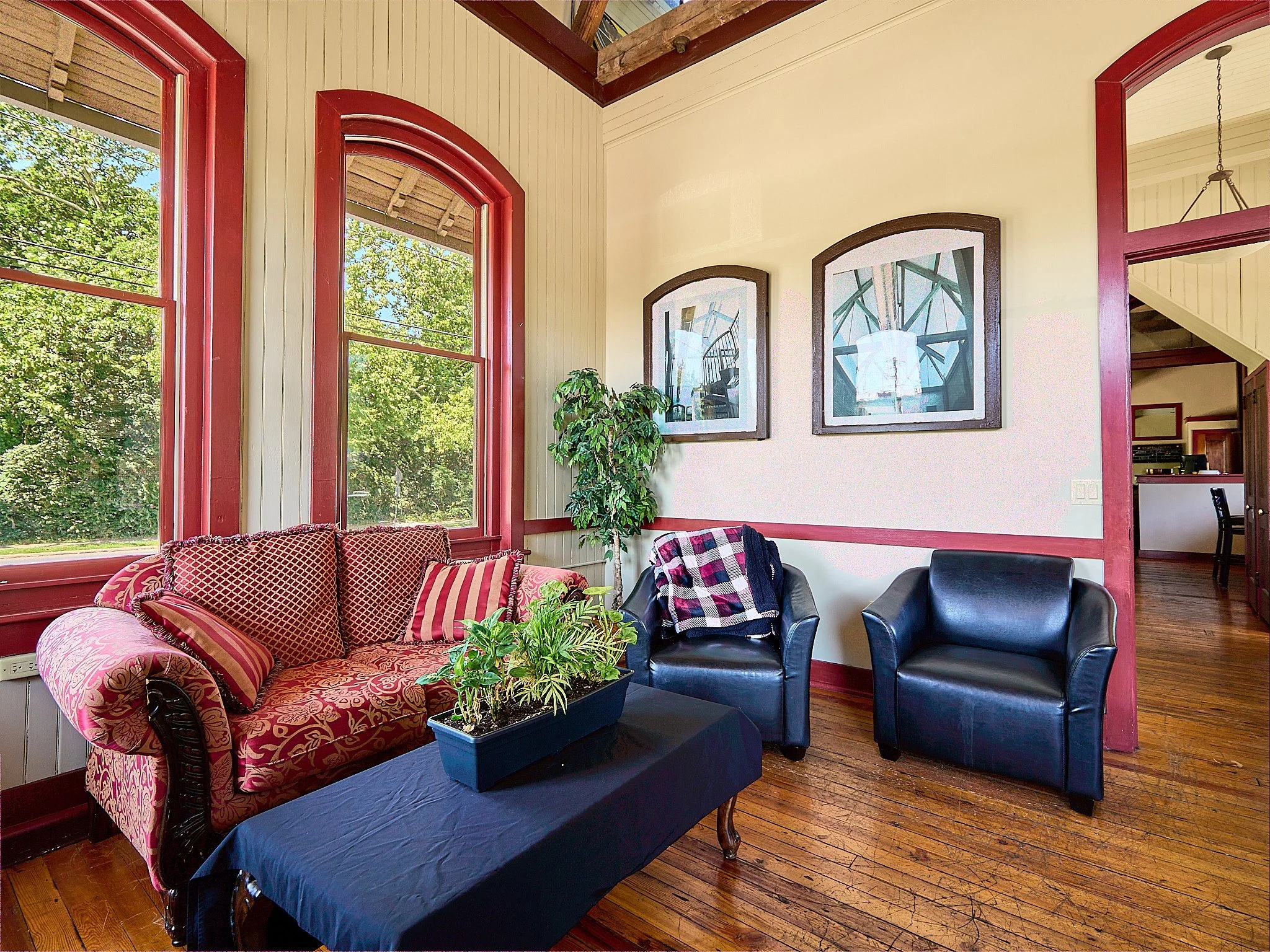 Cozy living room with large windows, red trim, a patterned sofa with pillows, two black leather armchairs with a blanket, a tall green plant, and framed artwork on beige walls.