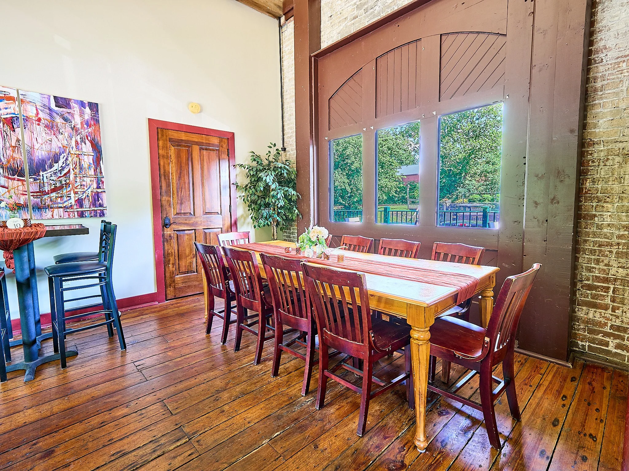 A dining room with a wooden table covered by a red tablecloth, surrounded by wooden chairs, next to brick and wood walls, with large windows showing trees outside, and a plant near a wooden door.