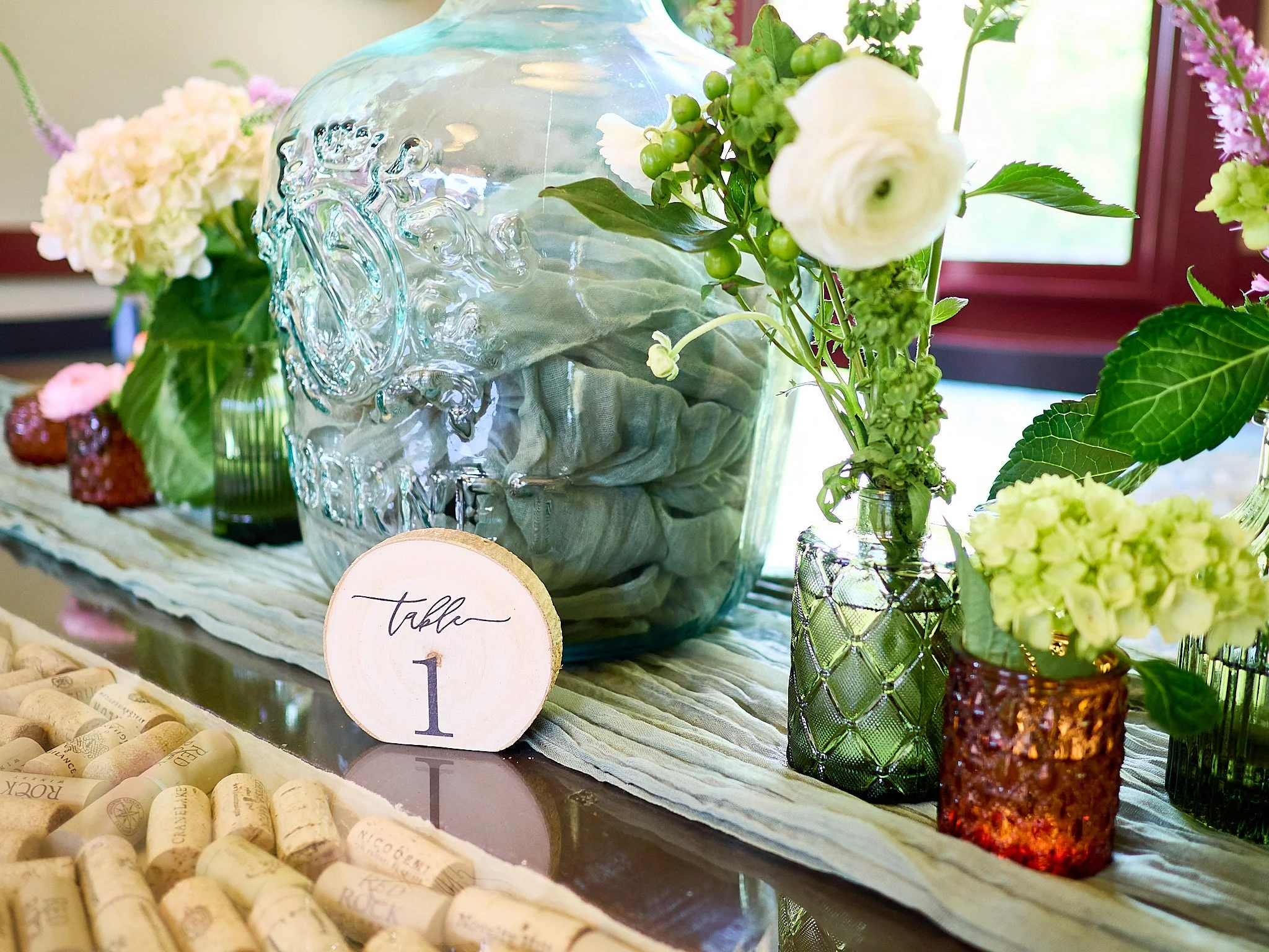 Decorative table centerpiece featuring glass vases with white and green flowers, a wooden table number sign marked "Table 1," and wine corks scattered at the front.