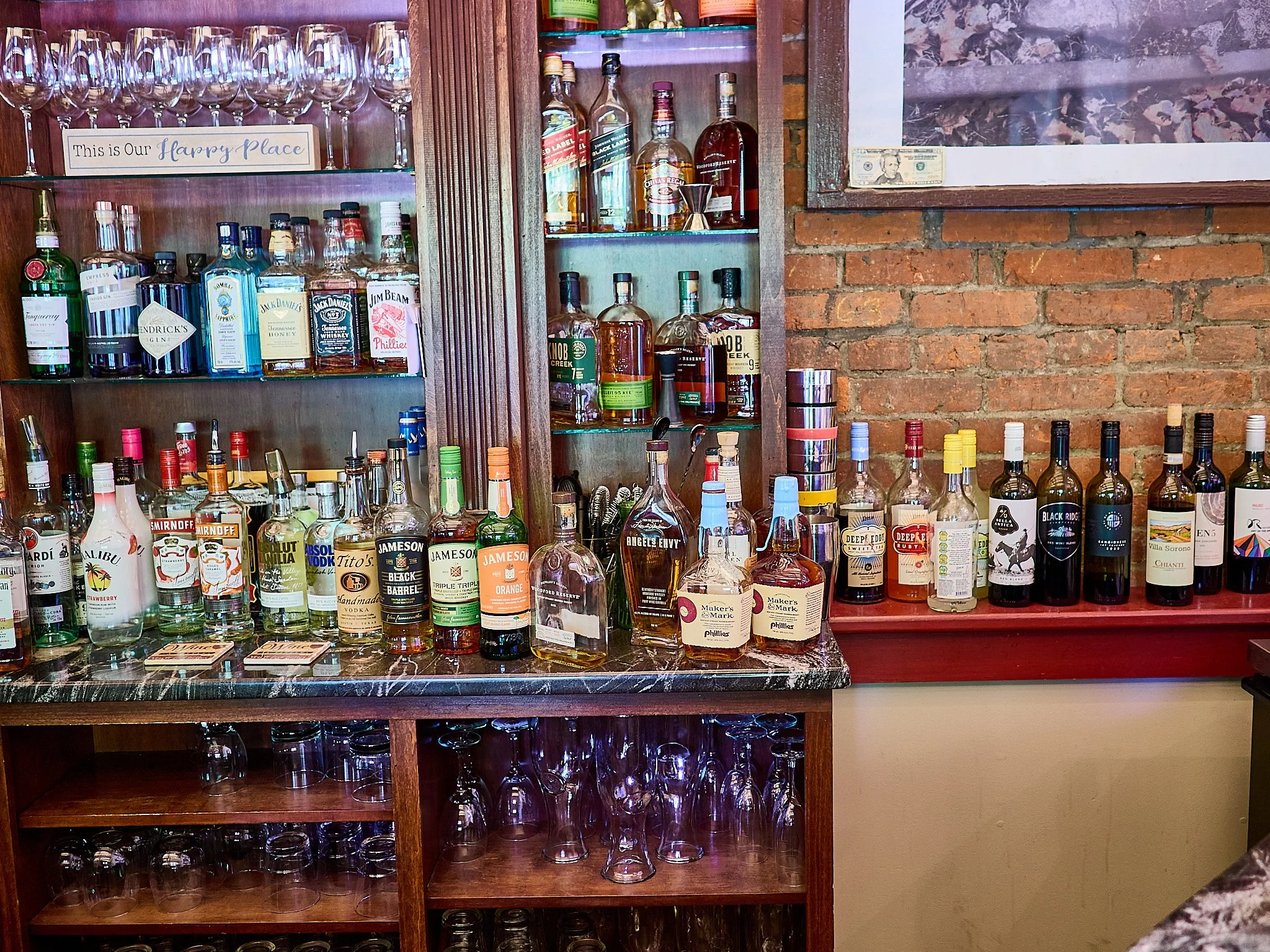 A bar counter with various bottles of alcohol including whiskey and vodka, glassware on shelves and under the counter, and a brick wall background.