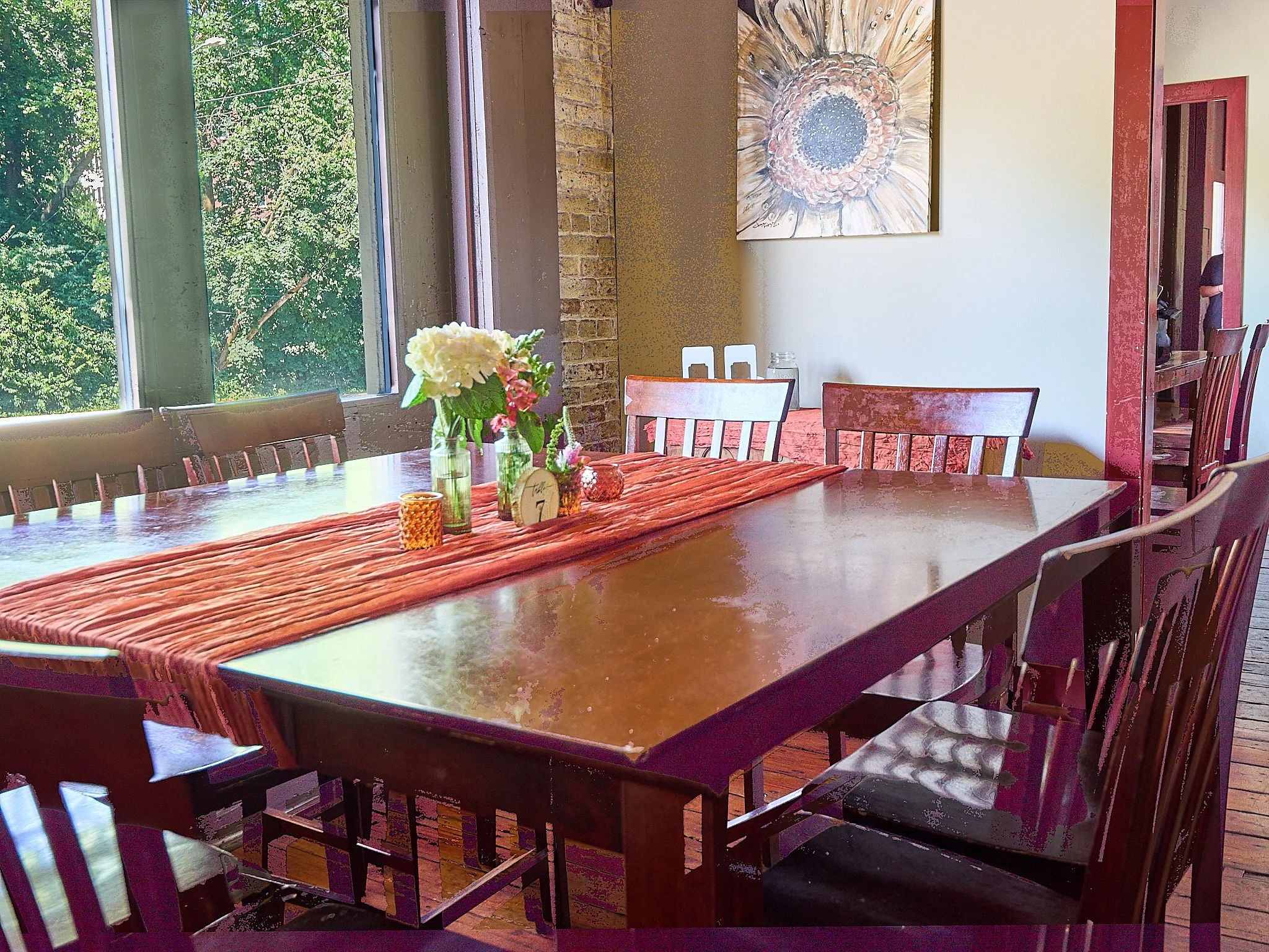 Dining room with large wooden table, surrounded by mismatched wooden chairs, decorated with a orange table runner, glass vases with flowers, and candles, next to large windows with green trees outside, and a wall with a sunflower painting.