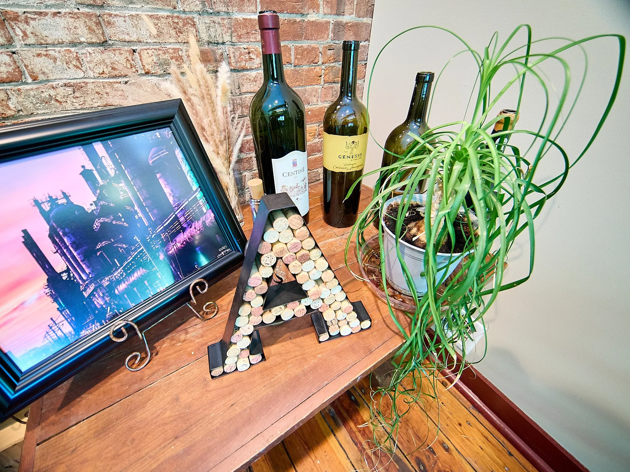 Decorative wooden table with a framed aircraft photo, an illuminated letter 'A' made of corks, three wine bottles, a potted green plant with long leaves, and a small decorative black object, all set against a brick wall and wooden floor.