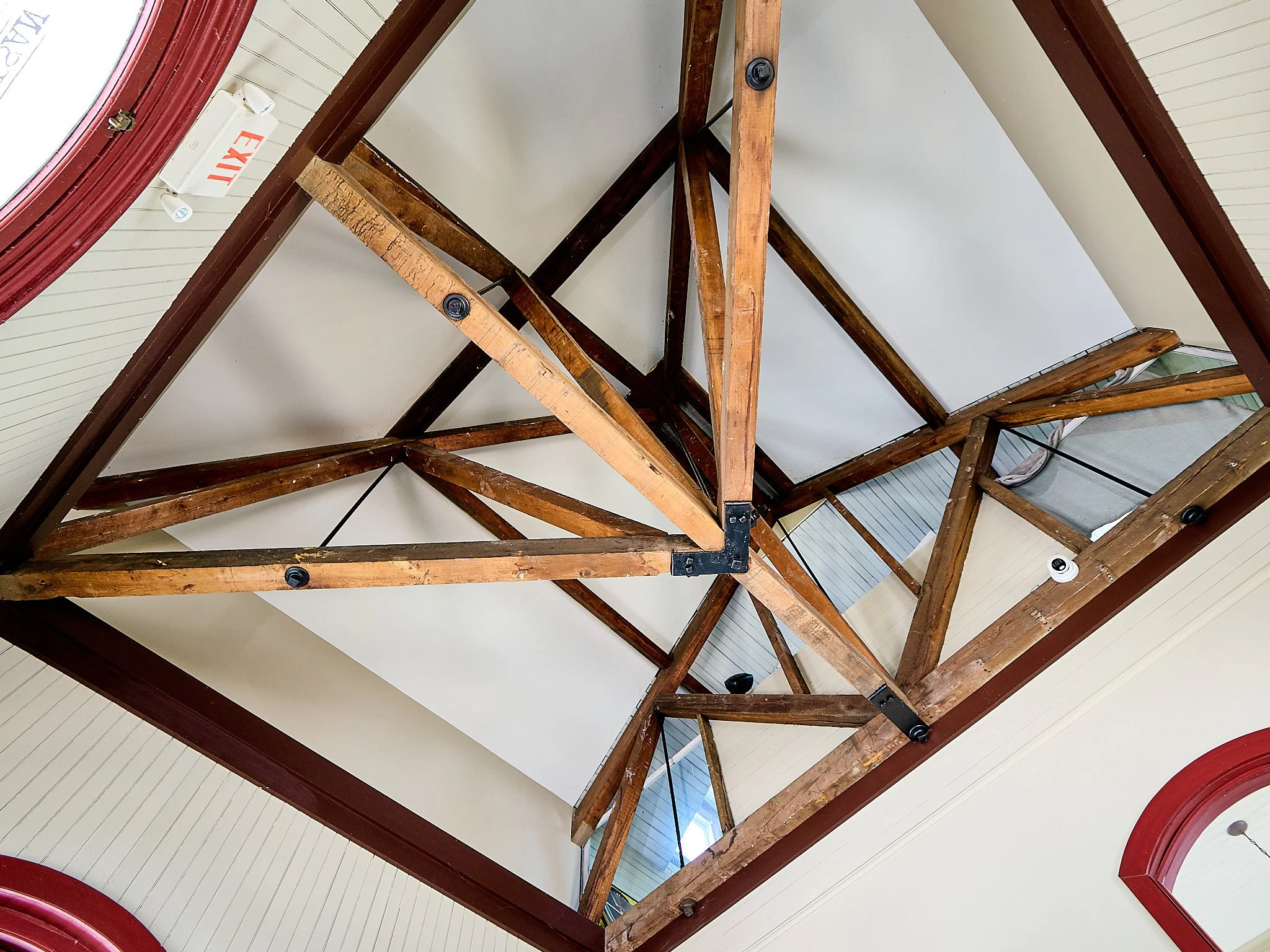 View of the ceiling showing wooden beams and framework, with a white wall and part of a red window frame visible.