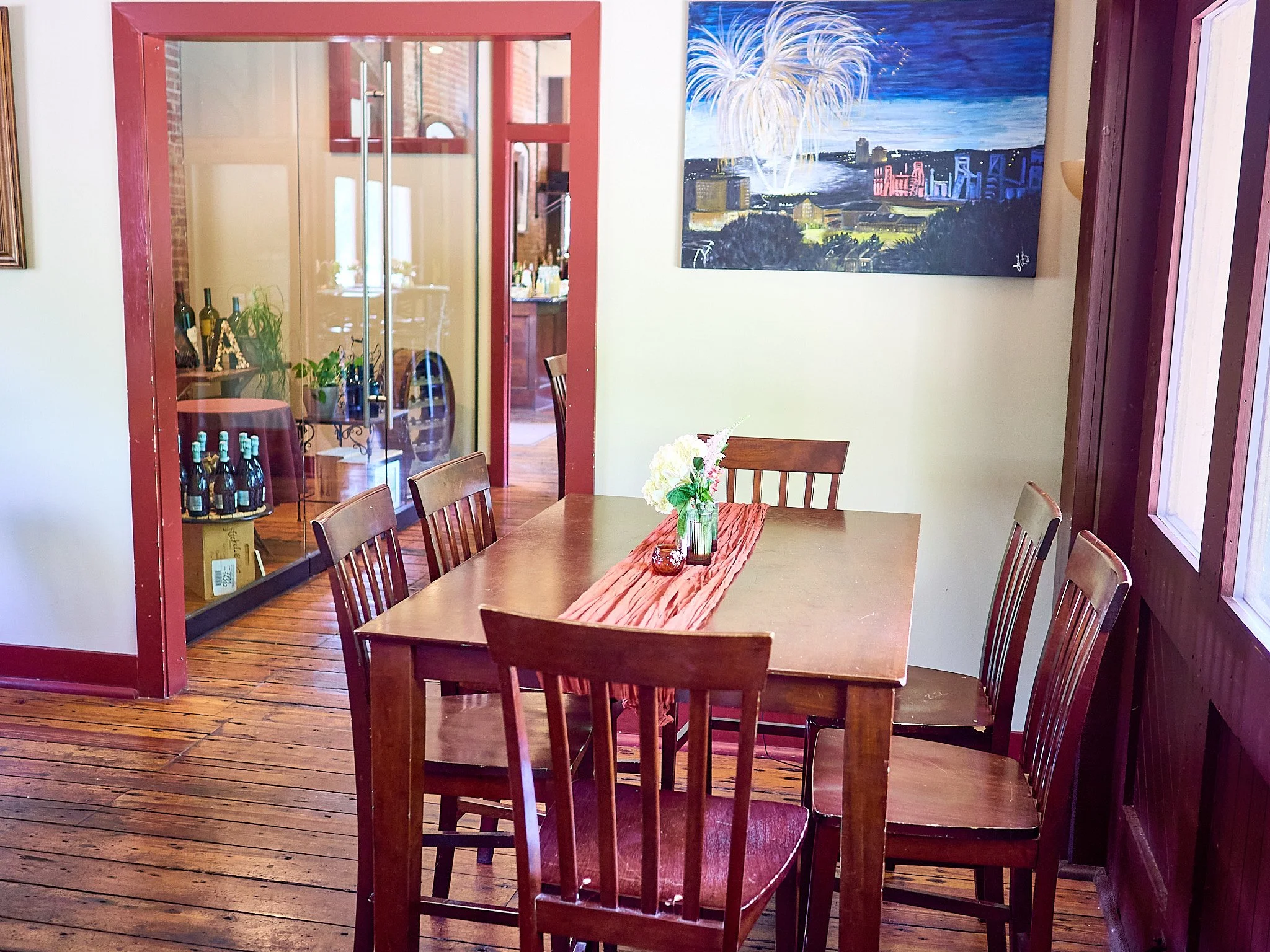 A dining room with a wooden table surrounded by six wooden chairs, a vase of flowers on a fabric table runner, a colorful cityscape and fireworks painting on the wall, and a glass-paneled door leading to another room with a bar area.