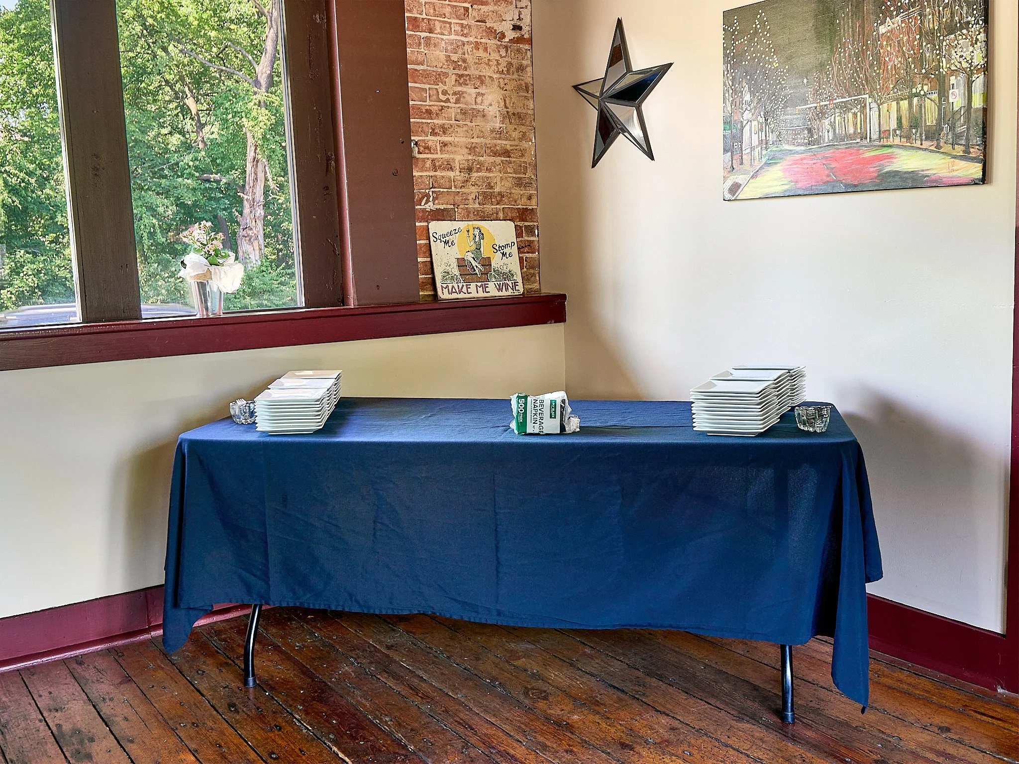 Table covered with a navy blue tablecloth, set for a gathering with stacks of white plates and small glass bowls, situated indoors near a window with a view of green trees outside, decorated with wall art and a star-shaped mirror.