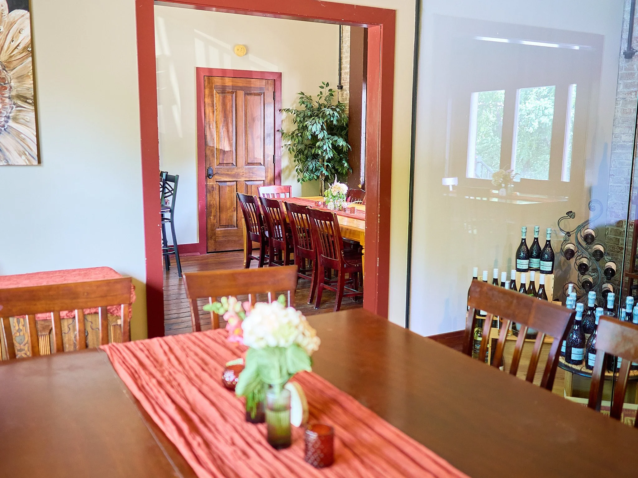A dining room with a wooden table and chairs, decorated with a floral centerpiece on an orange runner. Through a doorway, another room with multiple wooden chairs around a table and a large potted plant is visible. Shelves with wine bottles can be se