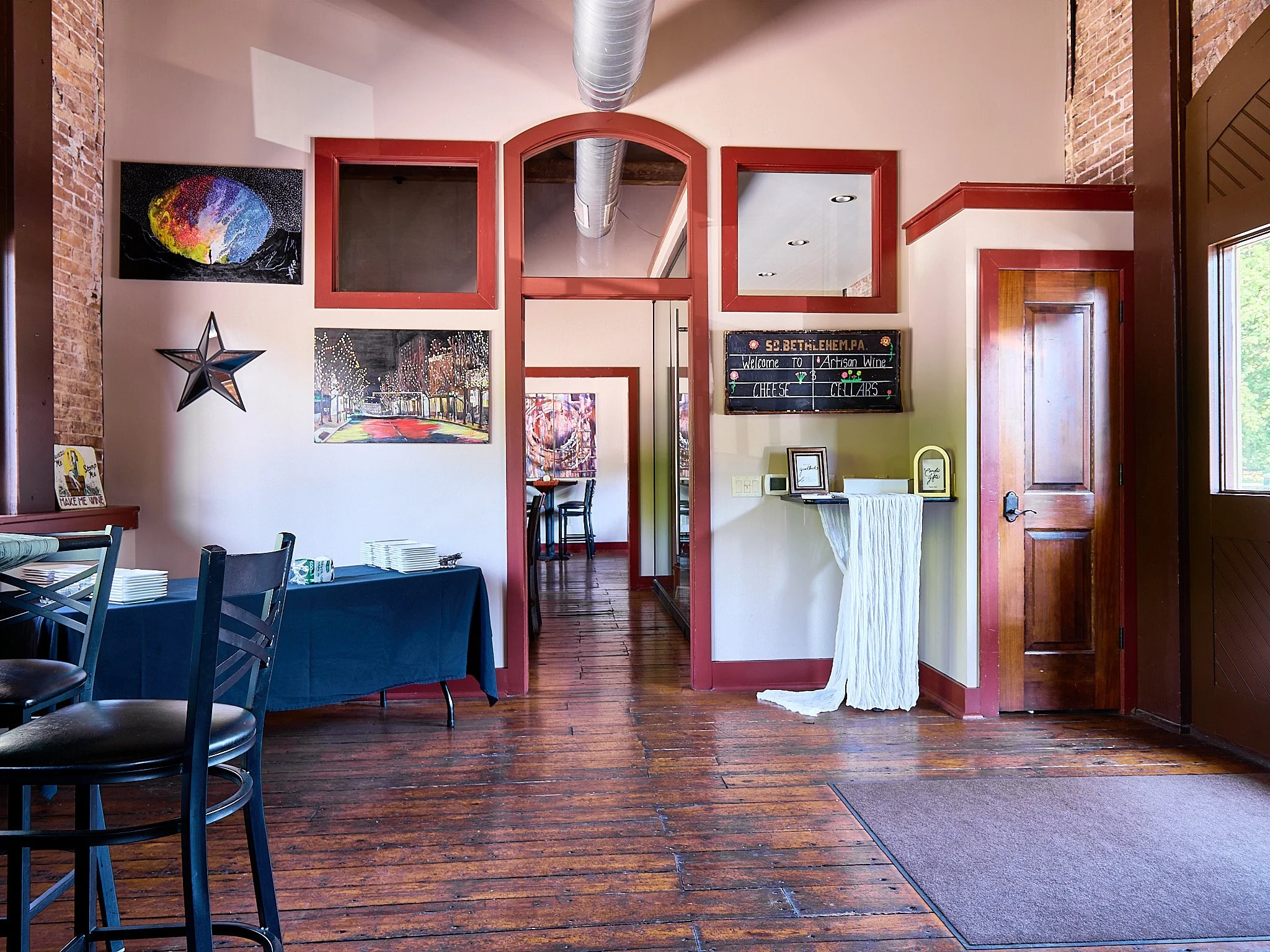 Interior of a cozy restaurant or cafe with wooden flooring, brick walls, and colorful artwork on the walls. There is a table with a blue tablecloth and chairs, and a small reception or checkout area near a door with a chalkboard sign.