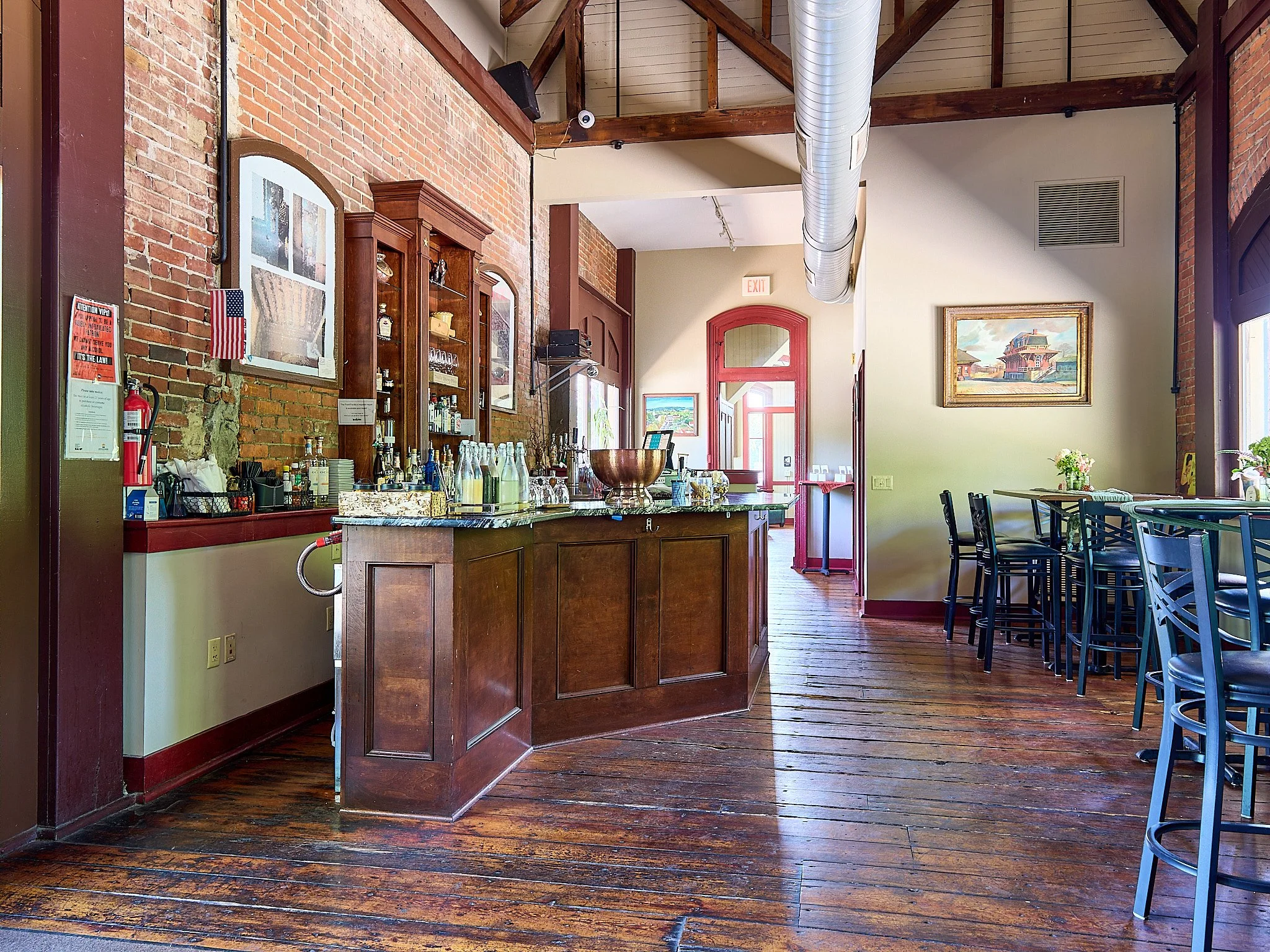 Interior of a rustic cafe with a brick wall, wooden bar counter with bottles, glasses, and a large bowl, with high tables and chairs, wooden flooring, and framed artwork on the walls.