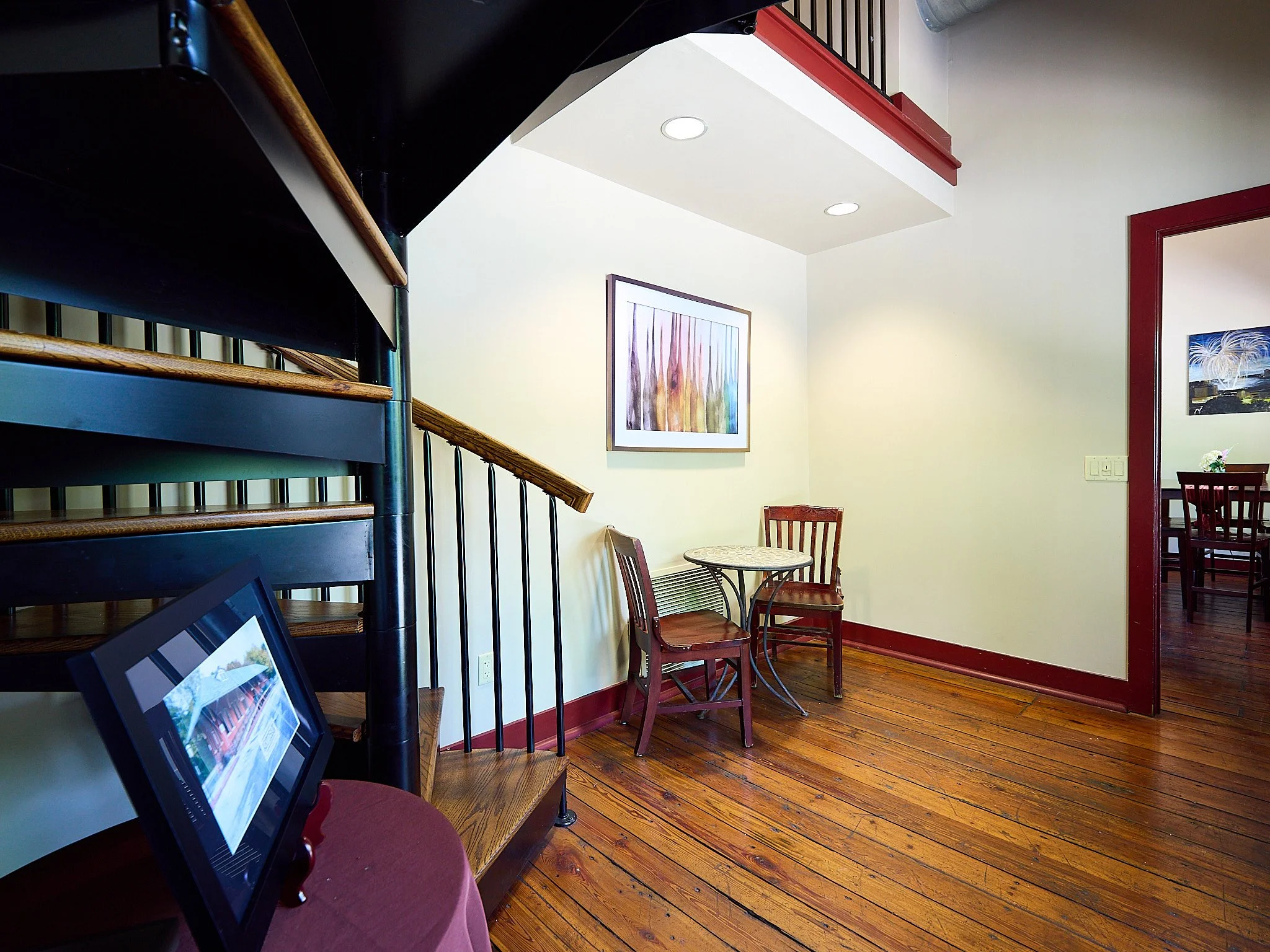 Interior view of a cozy dining or sitting area with a spiral staircase, a small round table with two chairs, a framed abstract art piece on the wall, and a doorway leading to another dining space decorated with flowers.