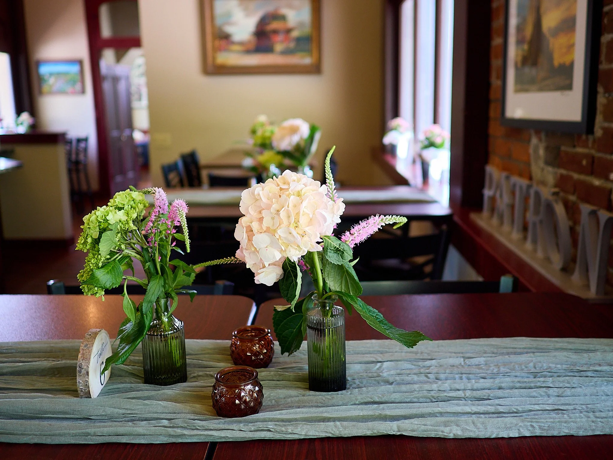 A dining table with a green table runner, decorated with three glass vases filled with pink and white hydrangeas and other flowers, set in a cozy room with artwork on the walls and a brick accent wall.
