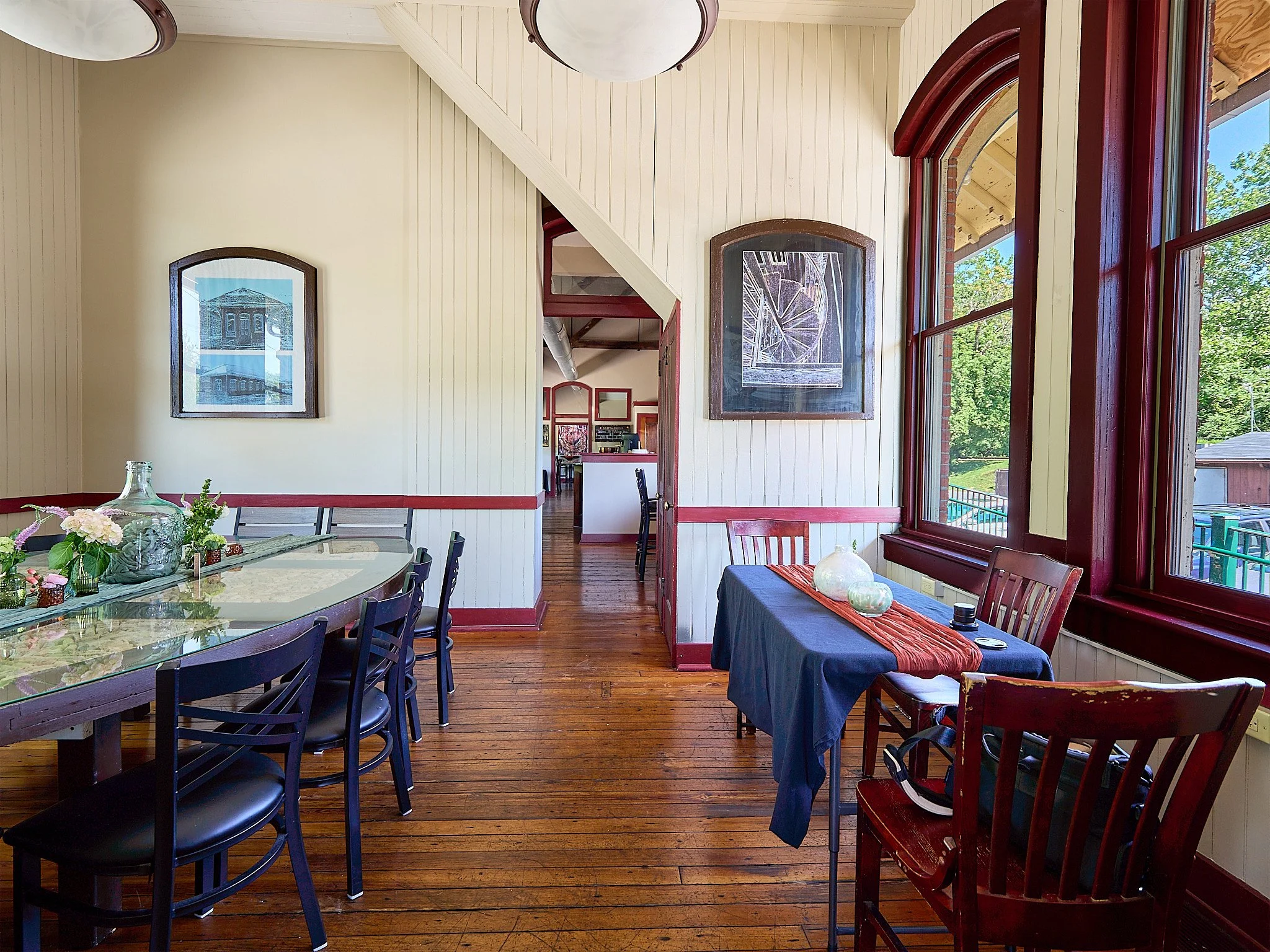 A cozy dining area with wooden floors, cream-colored paneled walls, and large windows. There is a round glass table with black chairs on the left and a small rectangular table with a dark tablecloth and wooden chairs on the right. The space is decora