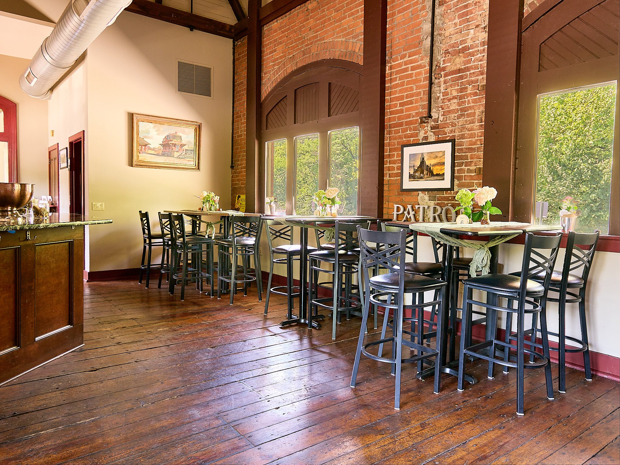 Interior of a restaurant with high wooden tables and black chairs, large windows letting in natural light, brick walls, and framed artwork, decorated with flowers and a 'Patron' sign.