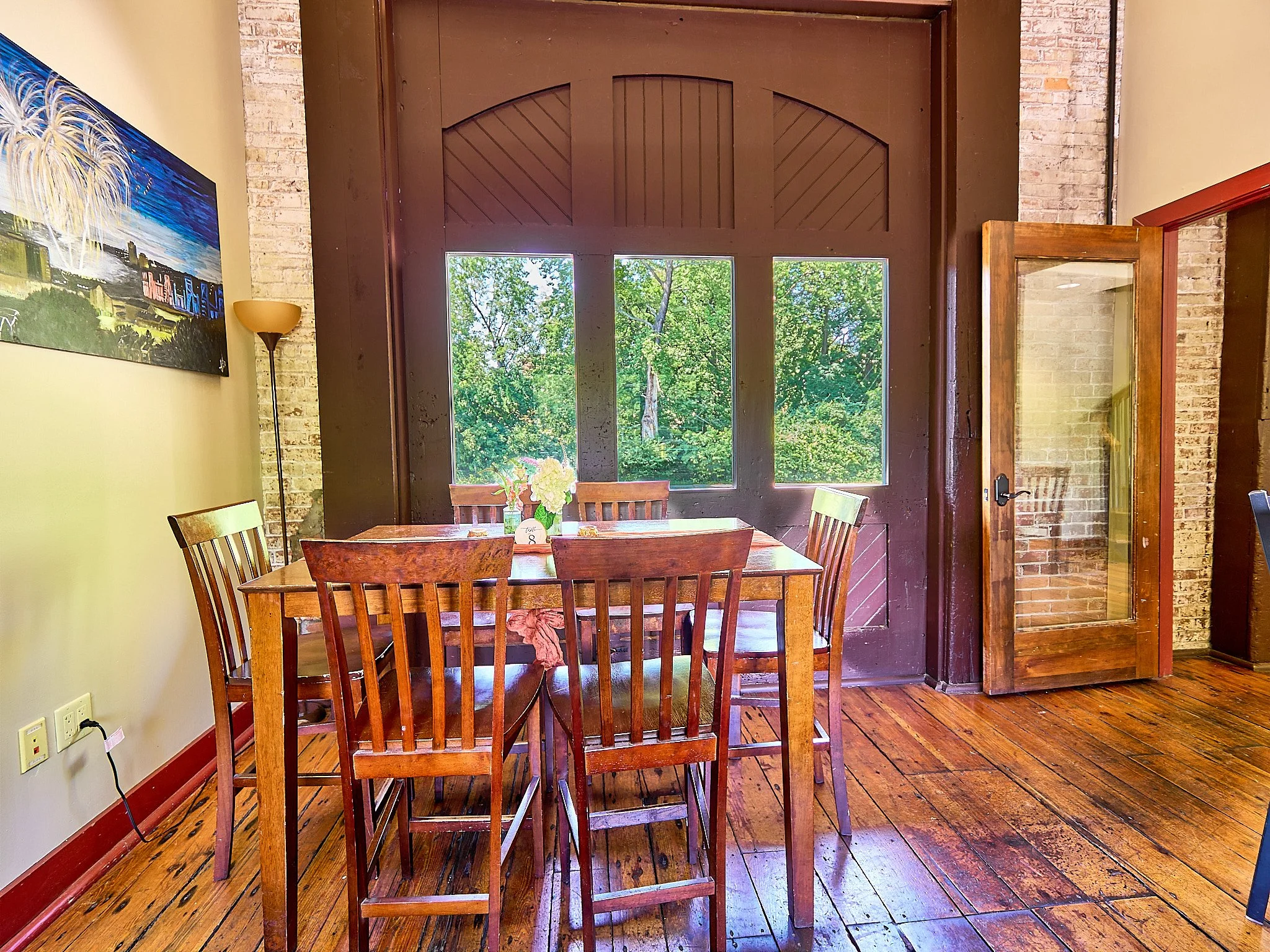 A dining area with a wooden table and chairs, large windows showing green trees outside, a brick wall, a wall painting, and a glass door.