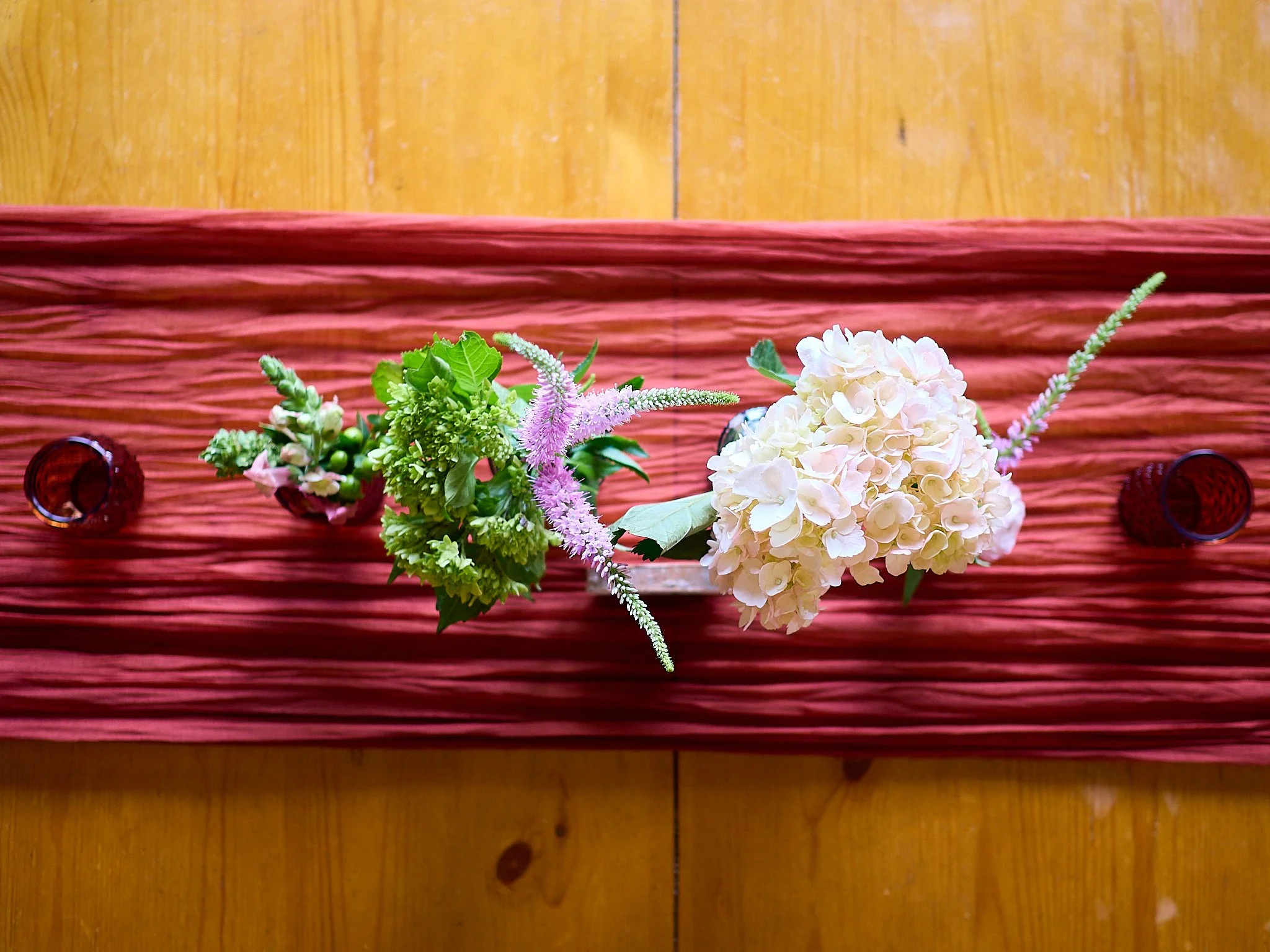 Top view of a floral centerpiece with white hydrangeas, pink and purple flowers, on a textured pink table runner on a wooden table.