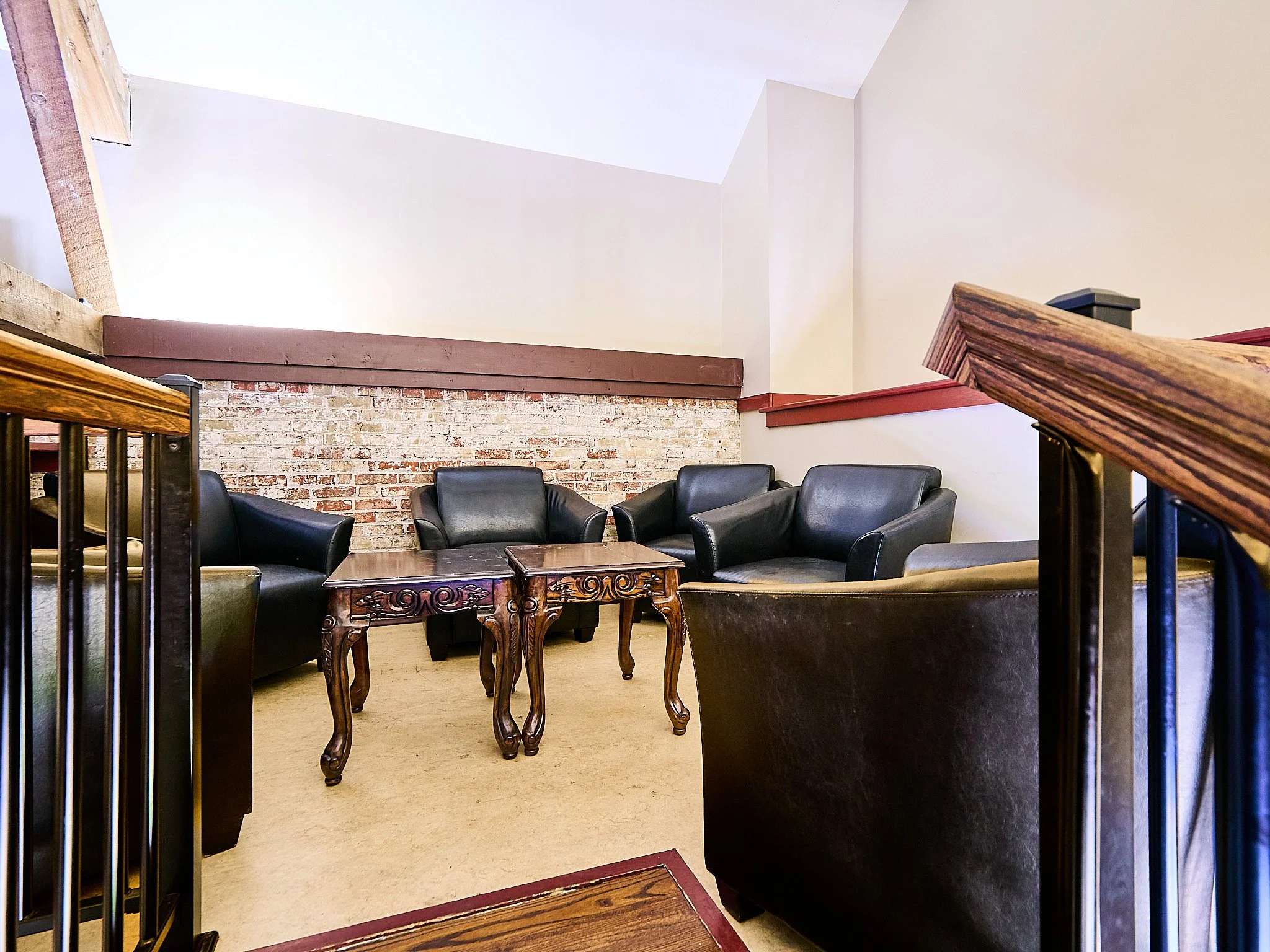 A cozy sitting area with four black leather armchairs arranged around a carved wooden coffee table in front of a brick accent wall, in a room with sloped ceilings and wooden stair railings.