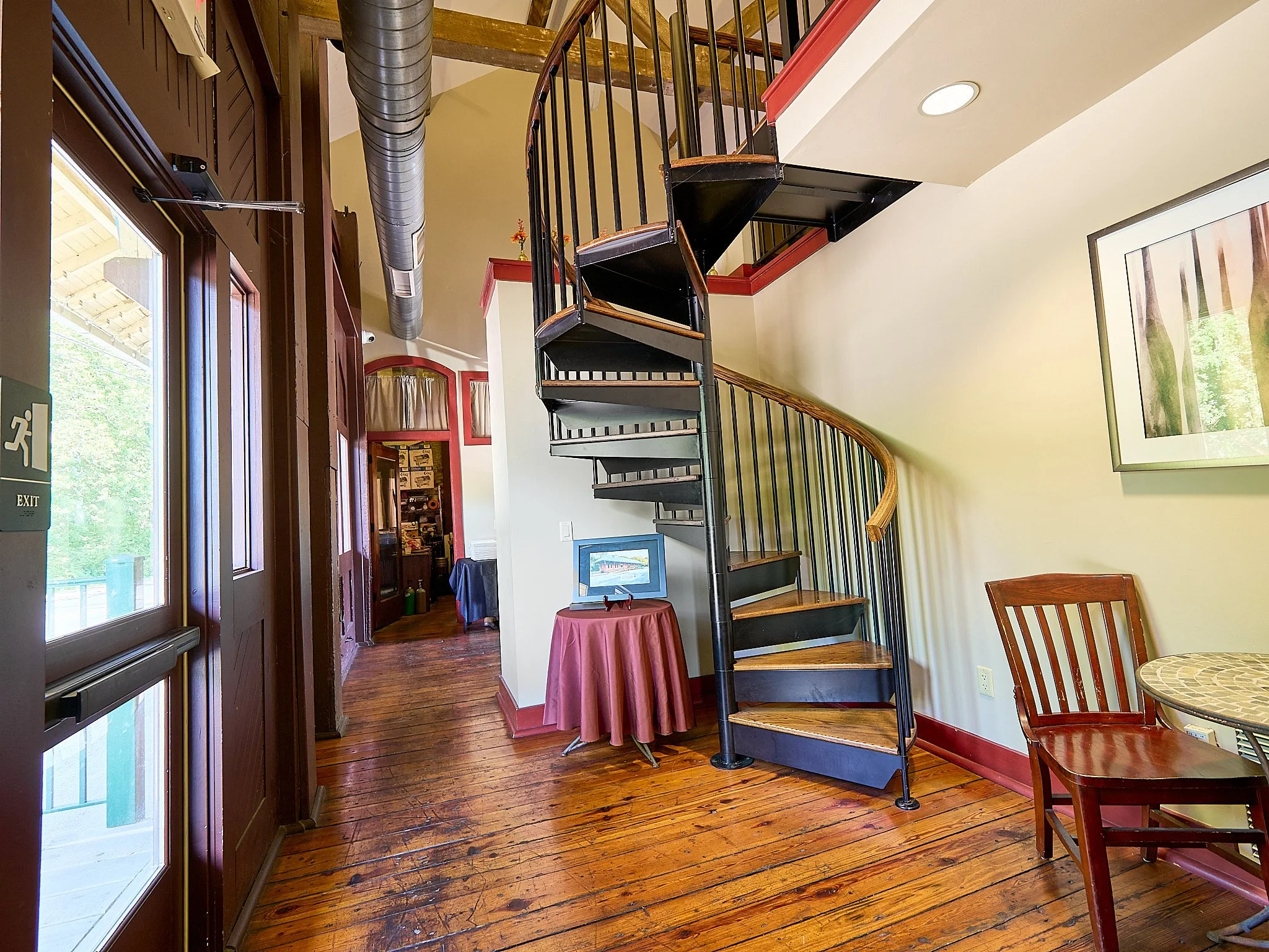 Interior of a cozy restaurant or cafe with wooden flooring, a spiral staircase, and a small table with a framed picture on it.
