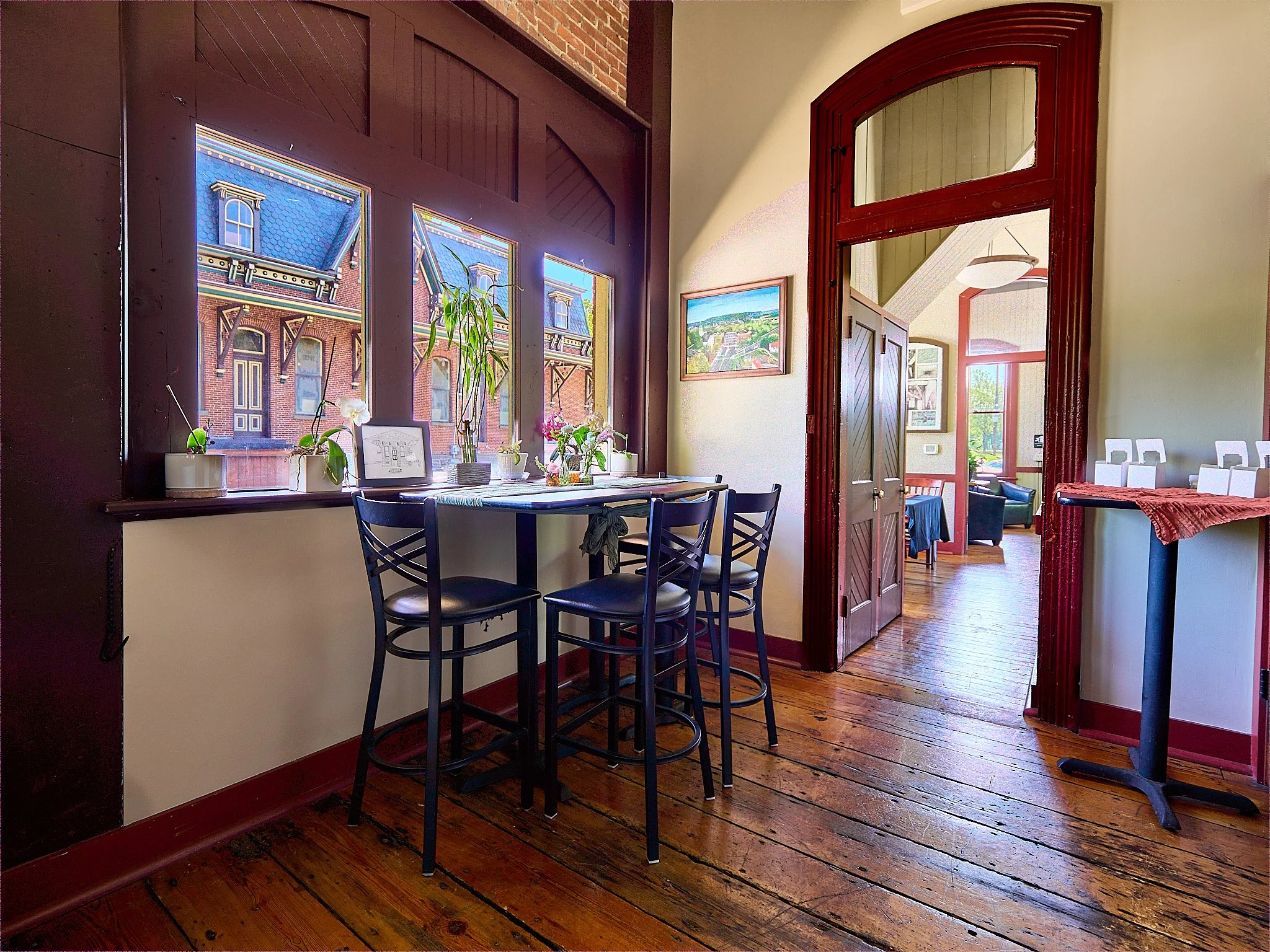 A cozy dining area with a high table and four black chairs next to a large window decorated with potted plants and framed pictures, with a view of city buildings outside.
