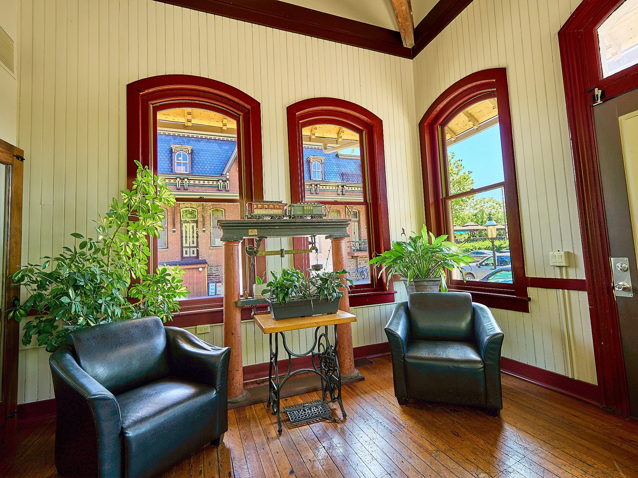 Interior of a room with wooden floors, cream-colored walls, red trim around large arched windows, and black leather chairs. Green plants are placed near the windows.