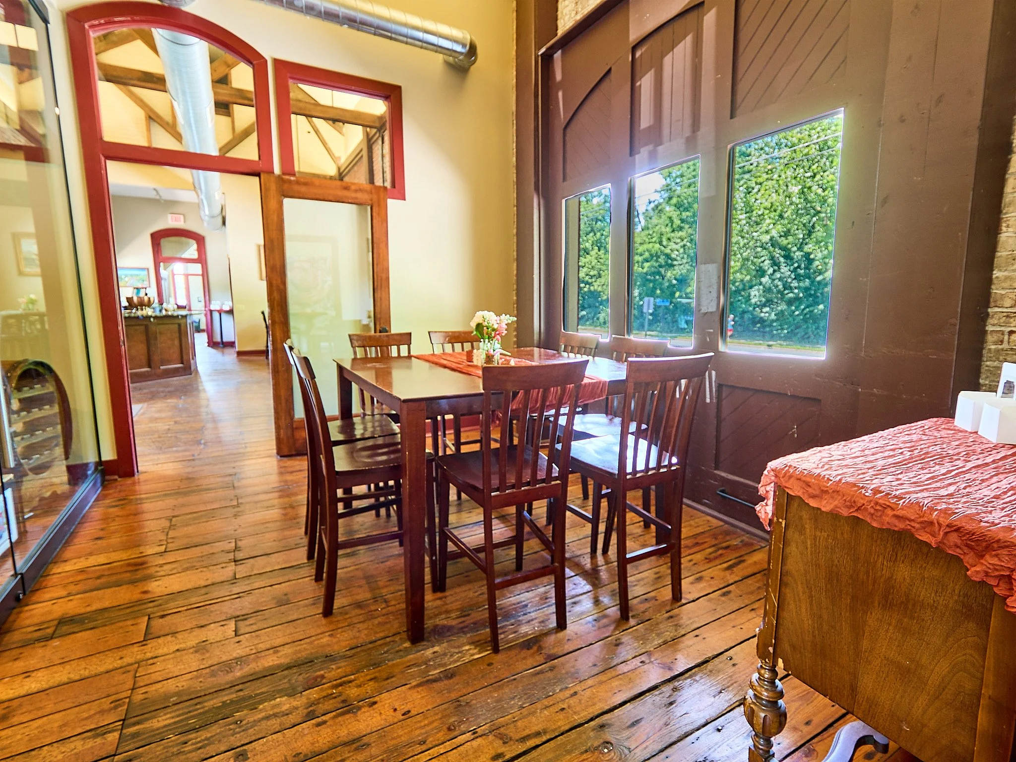 A cozy dining area with a wooden table and six matching chairs, decorated with a small flower arrangement, next to large windows showing green trees outside. The room has warm wooden floors and accents, with walls painted in soft colors and a side ta