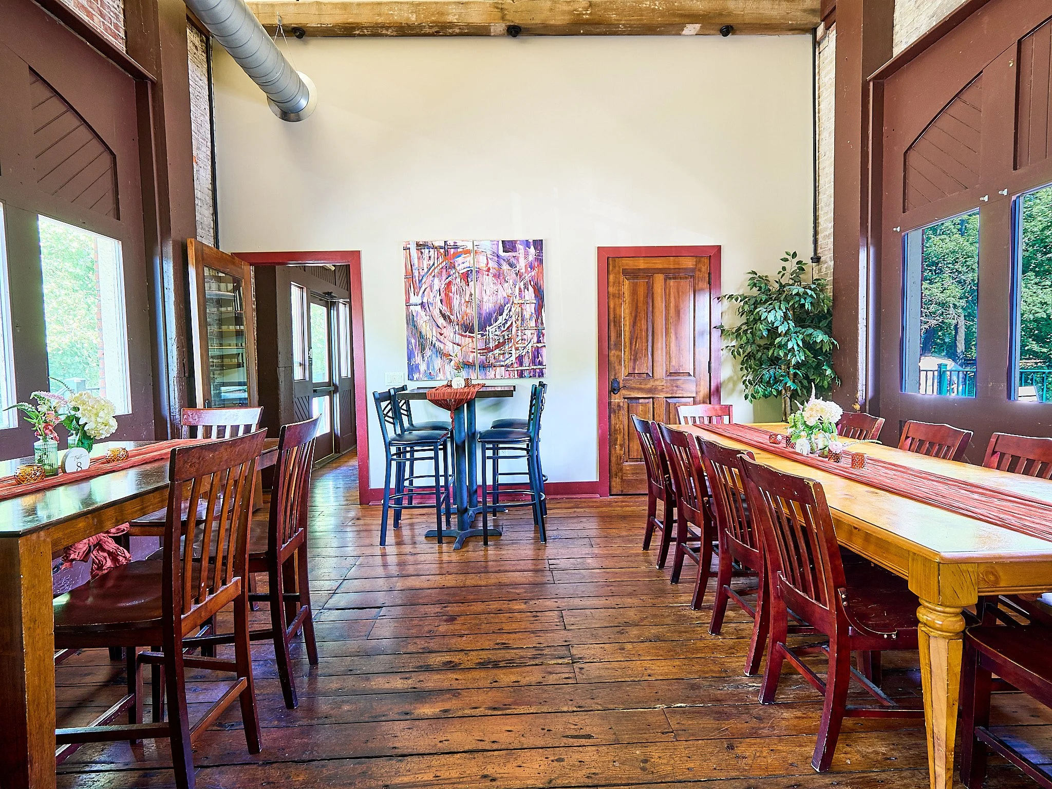 Interior of a dining room with wooden tables and chairs, decorated with vases of flowers, large windows showing trees outside, and a colorful abstract painting on the wall.