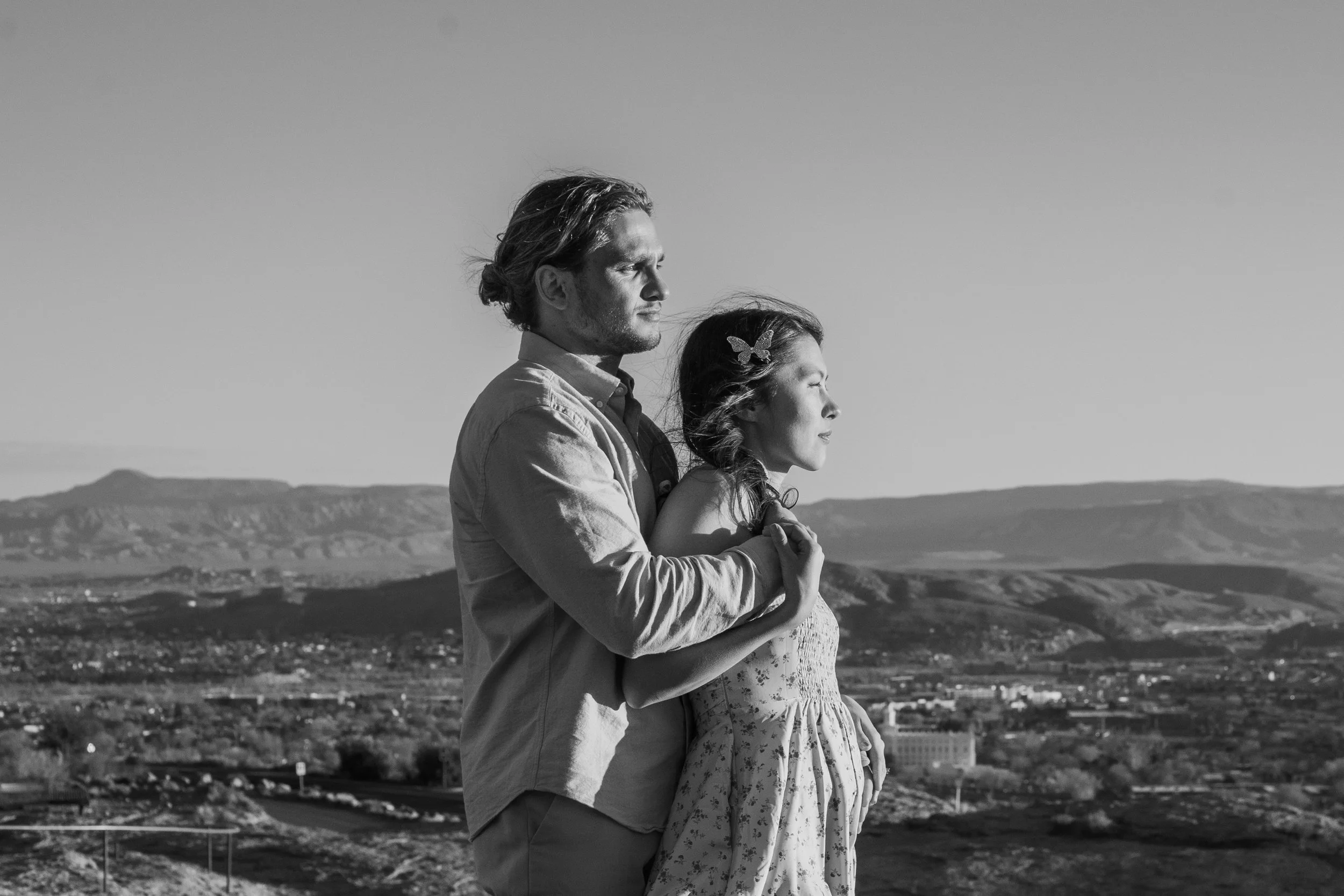 A black and white photo of a man and woman standing outdoors, with a landscape of mountains and a city in the background. The man is embracing the woman from behind, both with eyes closed, facing the right side of the image.