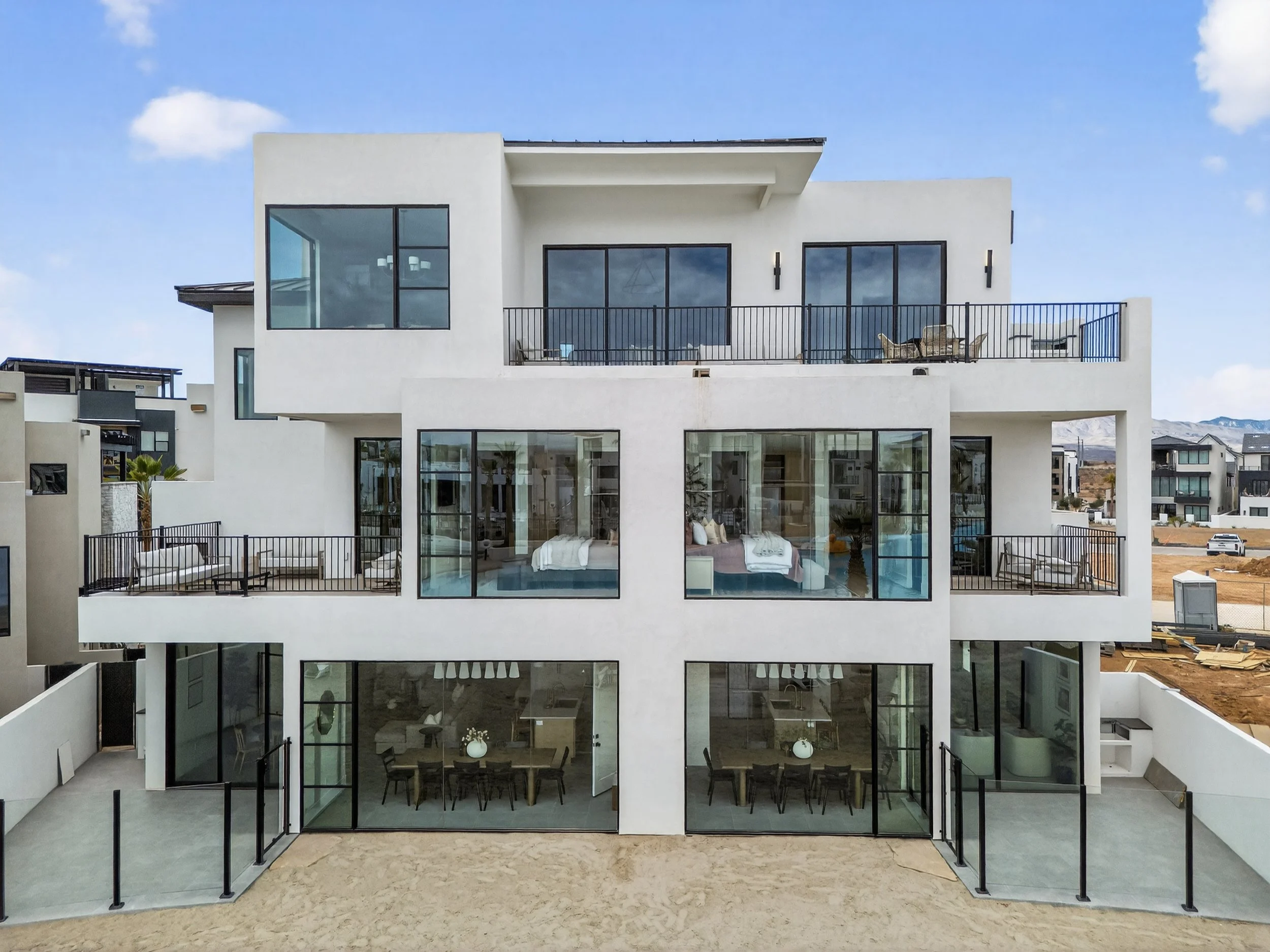 Modern multi-story white house with large glass windows and balconies, situated in a developing neighborhood with mountains in the background.